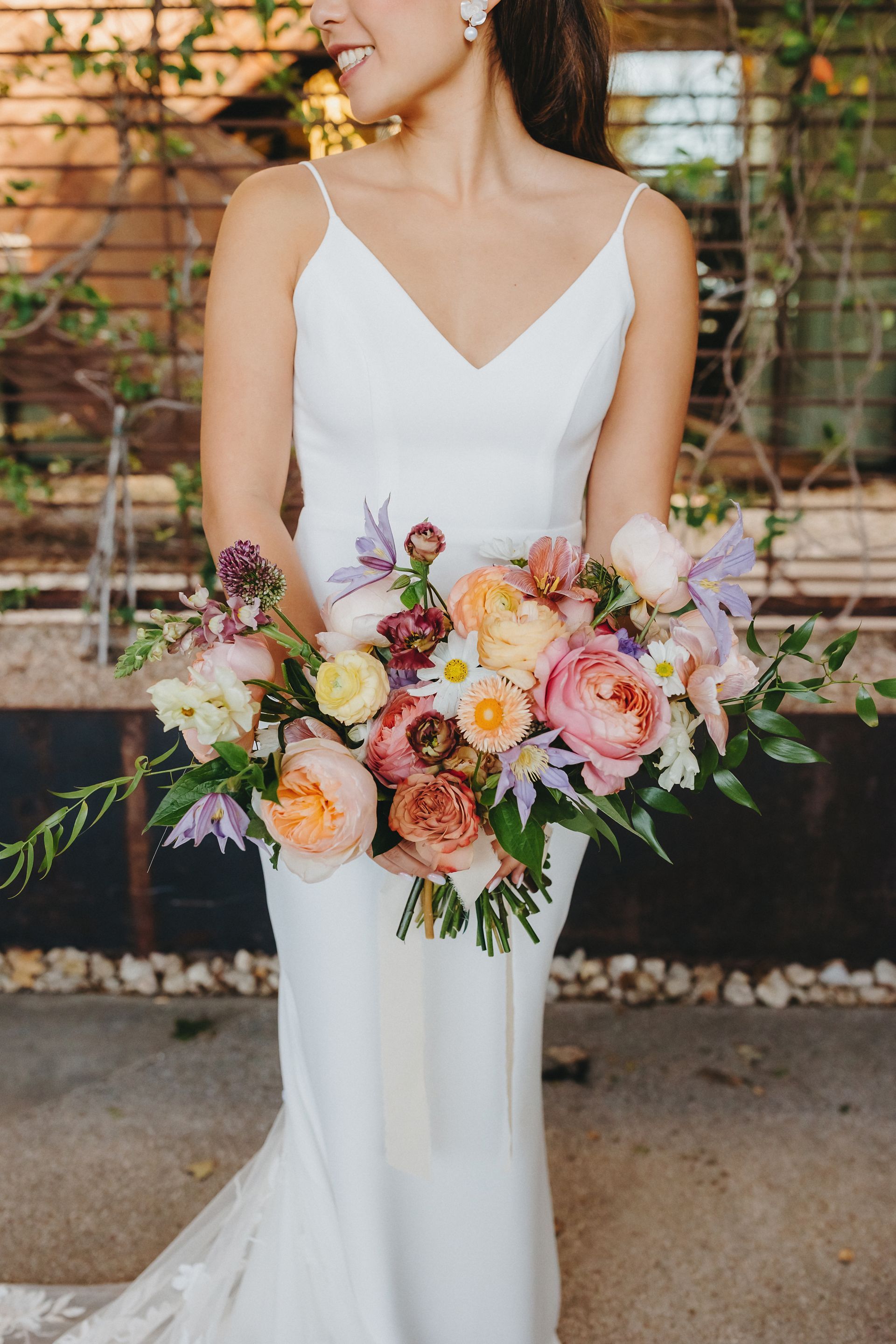 A person in a simple white V-neck wedding dress holds a large, colorful bouquet of garden roses and wildflowers.