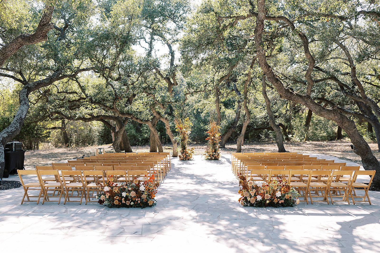 Rows of wooden folding chairs set up for an outdoor wedding ceremony under large, leafy oak trees with floral accents.