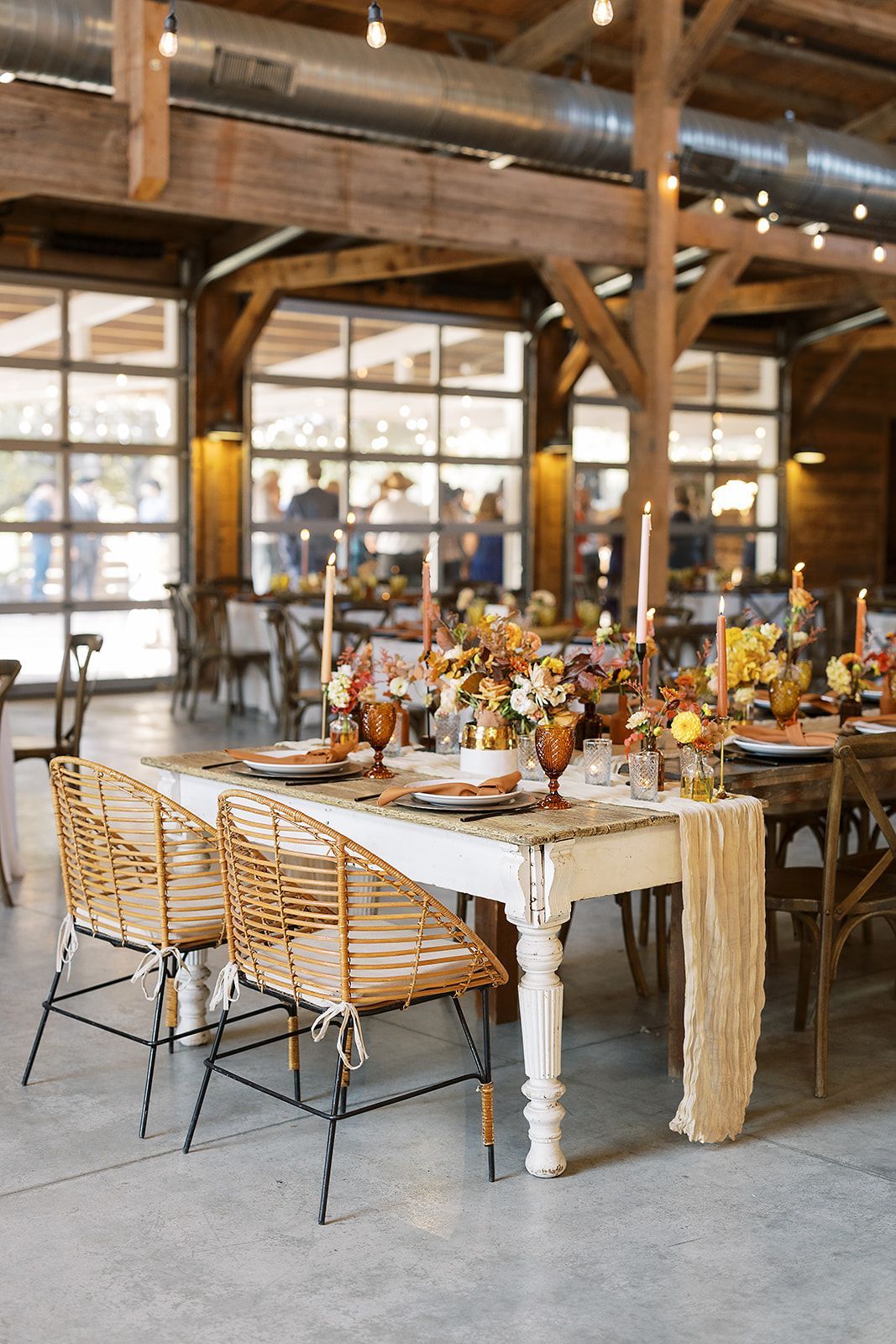 Rustic event space with a decorated dining table, wicker chairs, and fall-toned floral centerpieces in a barn setting.