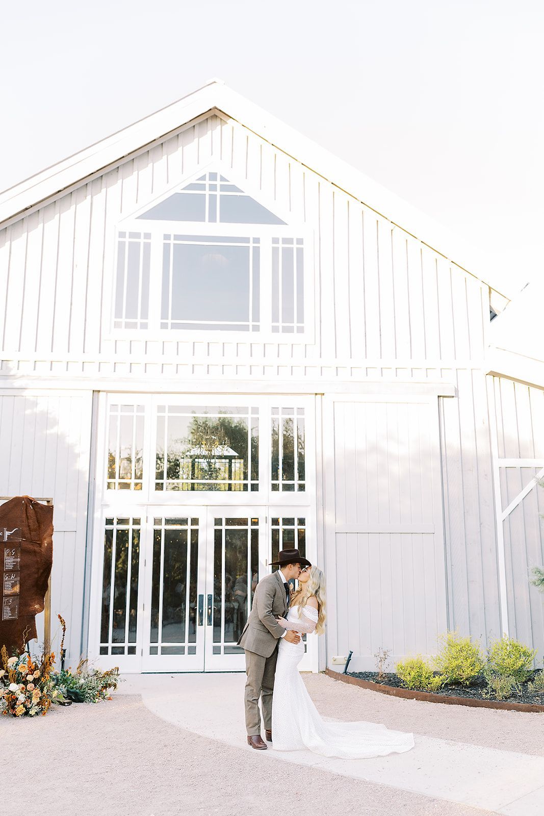 A couple in wedding attire kisses in front of a white barn with large glass doors on a gravel path.