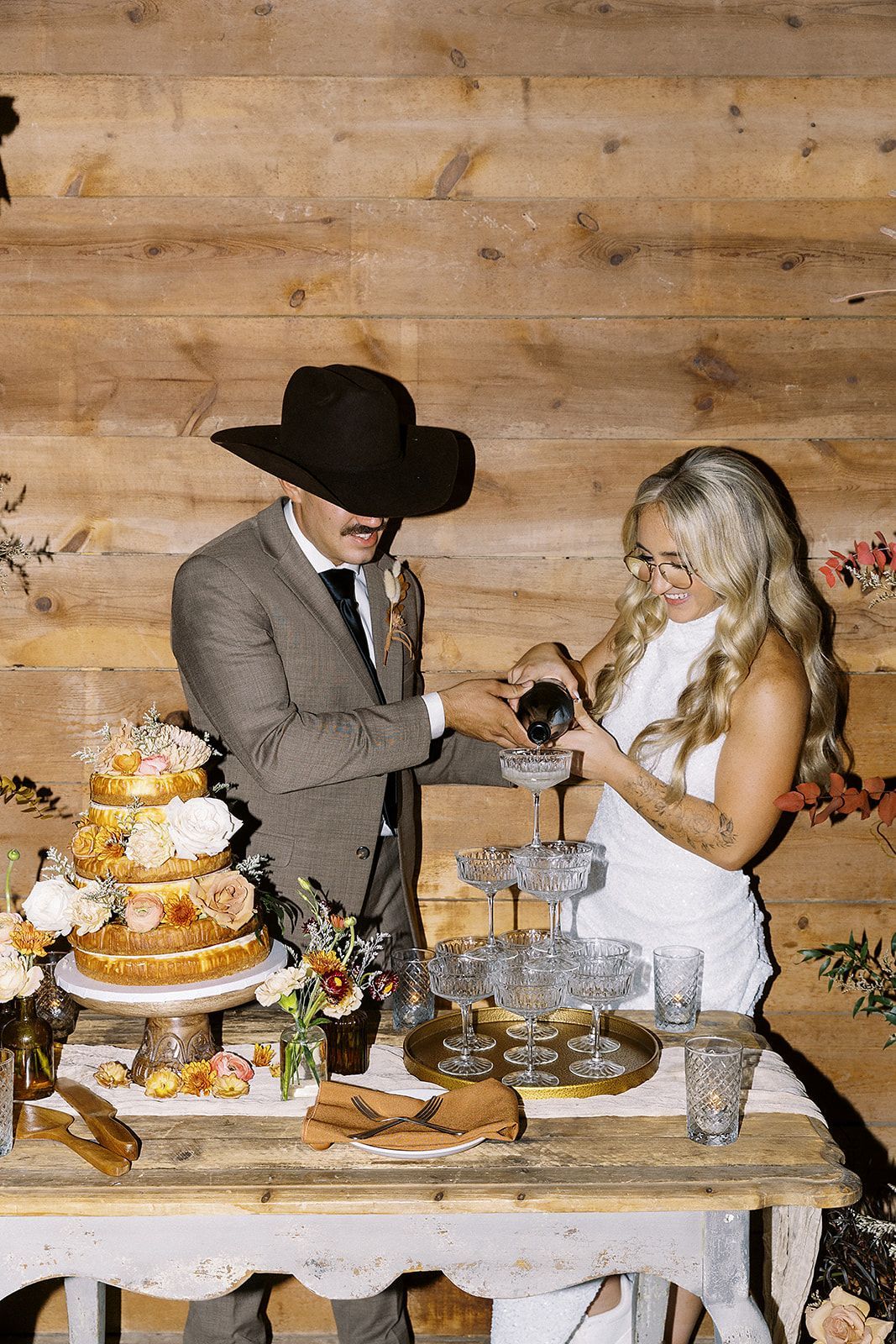 A person in a suit and cowboy hat and another in a white dress pour champagne into a stacked glass tower at a wedding.