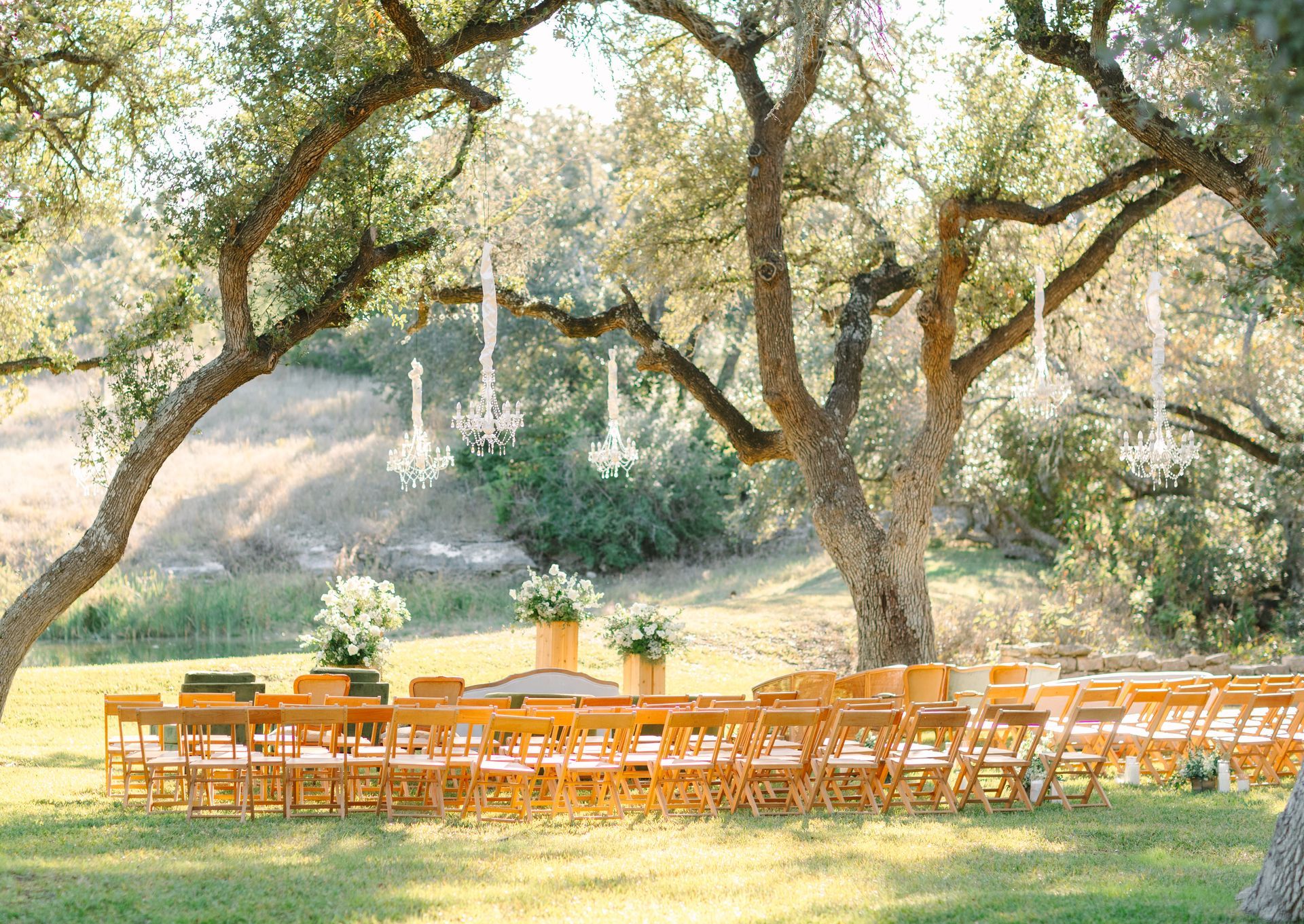 An outdoor wedding setup with rows of wooden chairs under large oak trees, decorated with hanging white chandeliers.