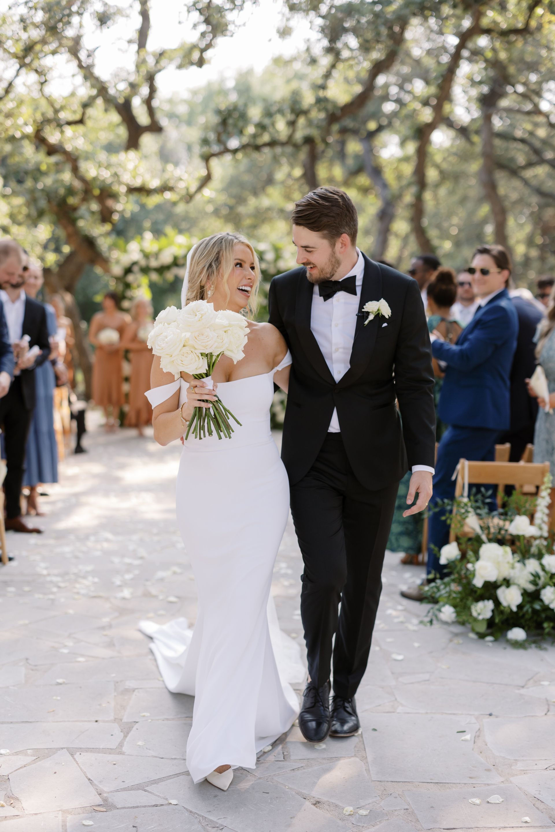 A smiling bride in a white wedding gown walks arm-in-arm with a groom in a black tuxedo during an outdoor wedding ceremony.