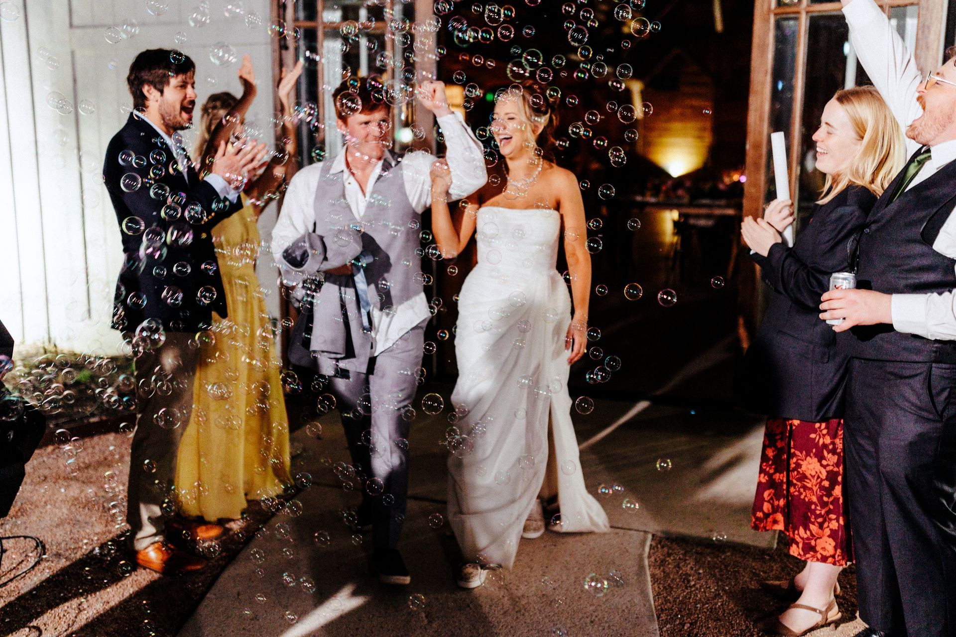 A bride and groom walk through a bubble-filled exit path, surrounded by cheering guests in evening attire.