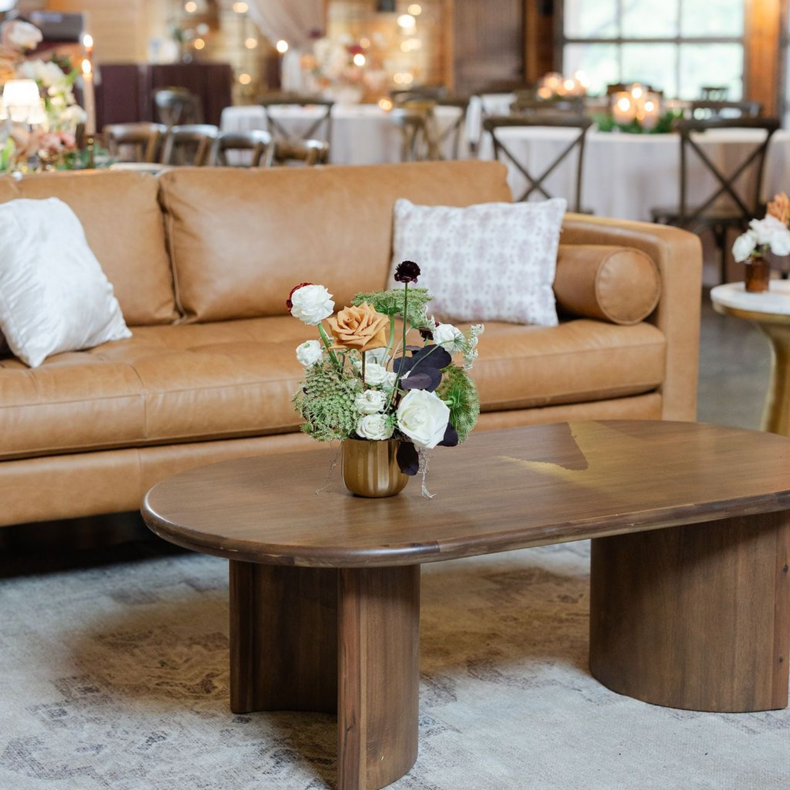 A tan leather sofa with pillows sits behind a wooden coffee table topped with a floral arrangement in The Addison Grove's venue.