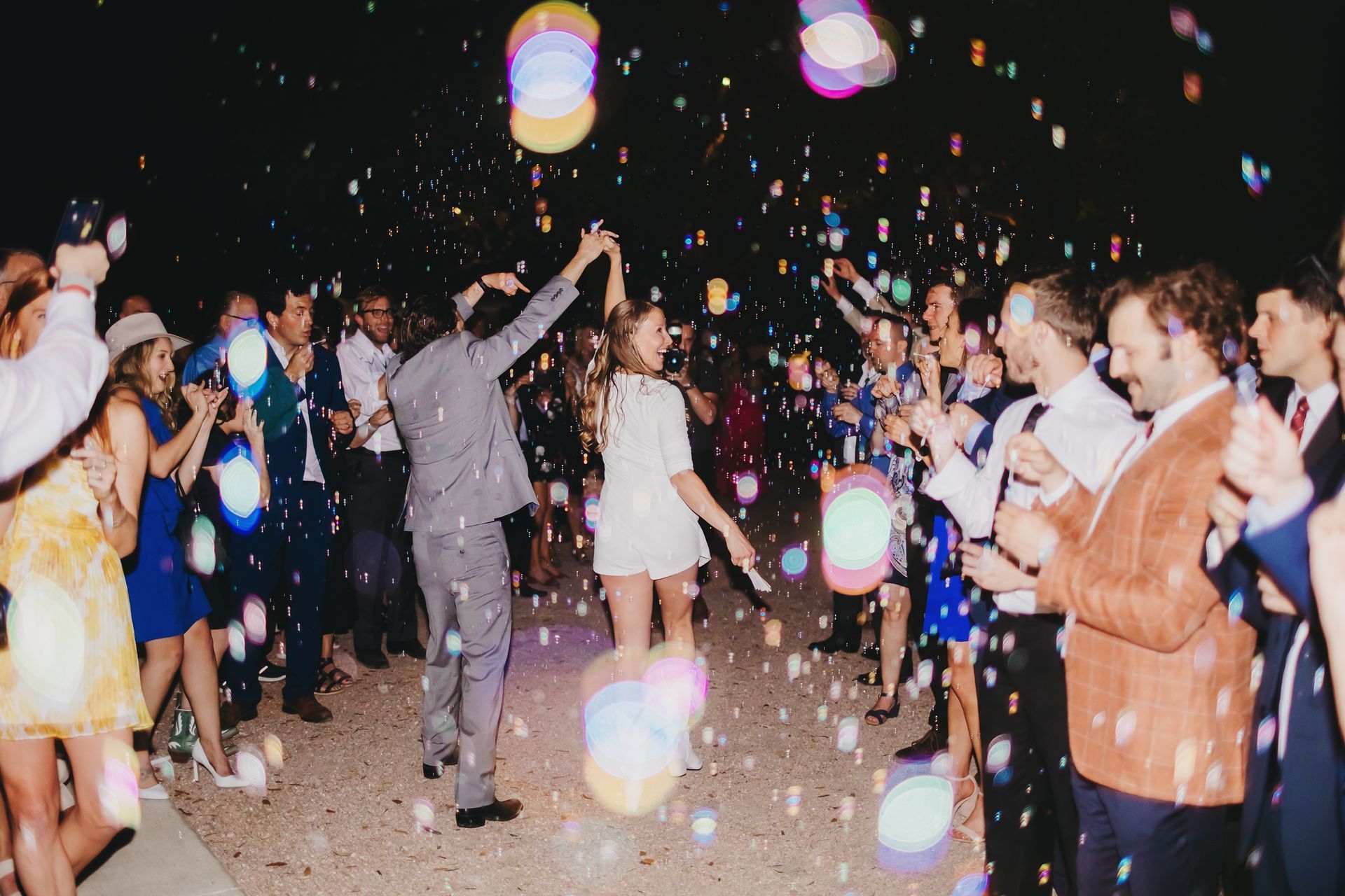 A newlywed couple dances through a crowd of guests blowing bubbles at a night celebration, illuminated by soft bokeh.