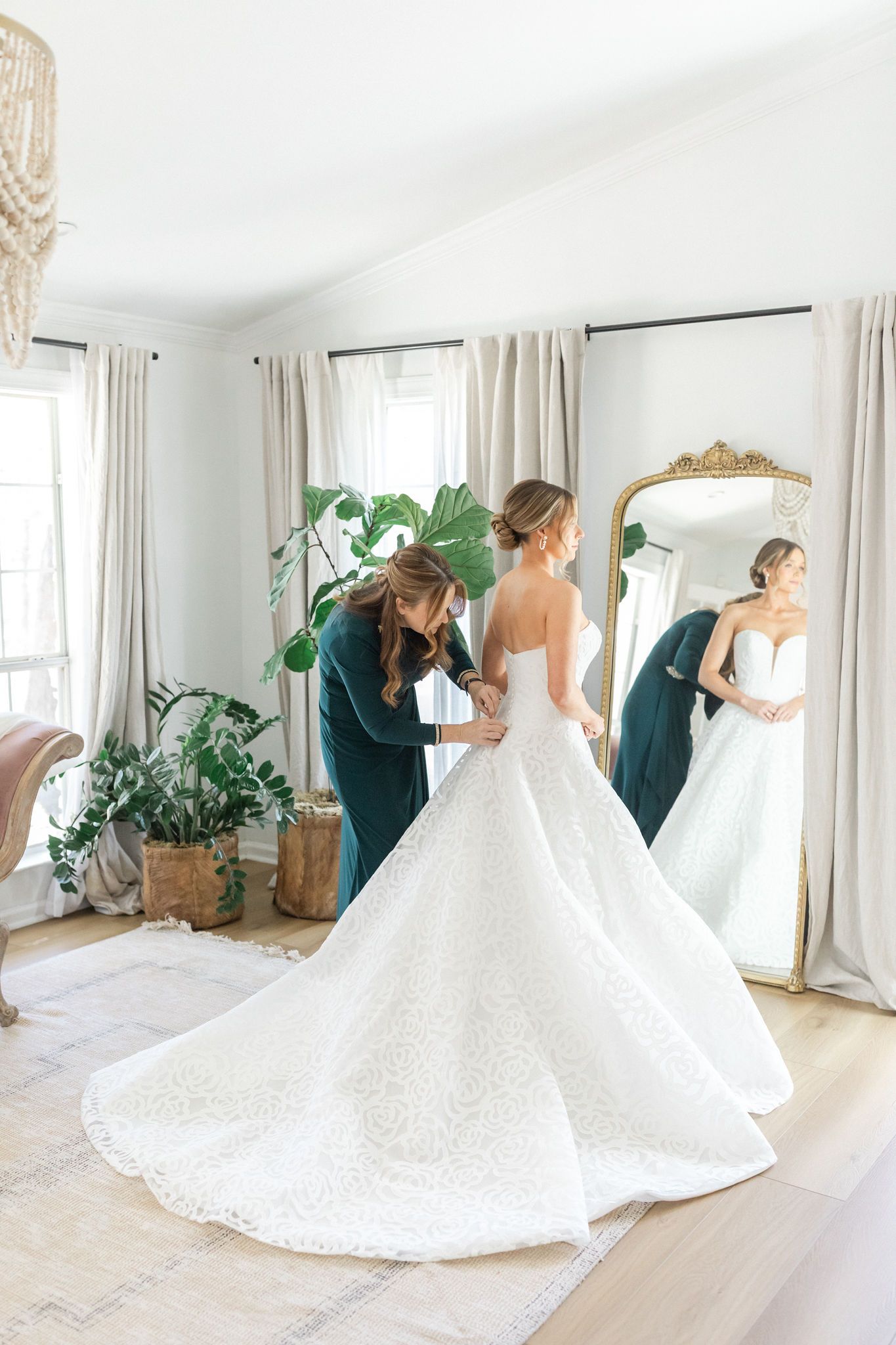 A person in a dark sweater helps a bride zip up her white, textured ballgown wedding dress in front of a floor mirror.