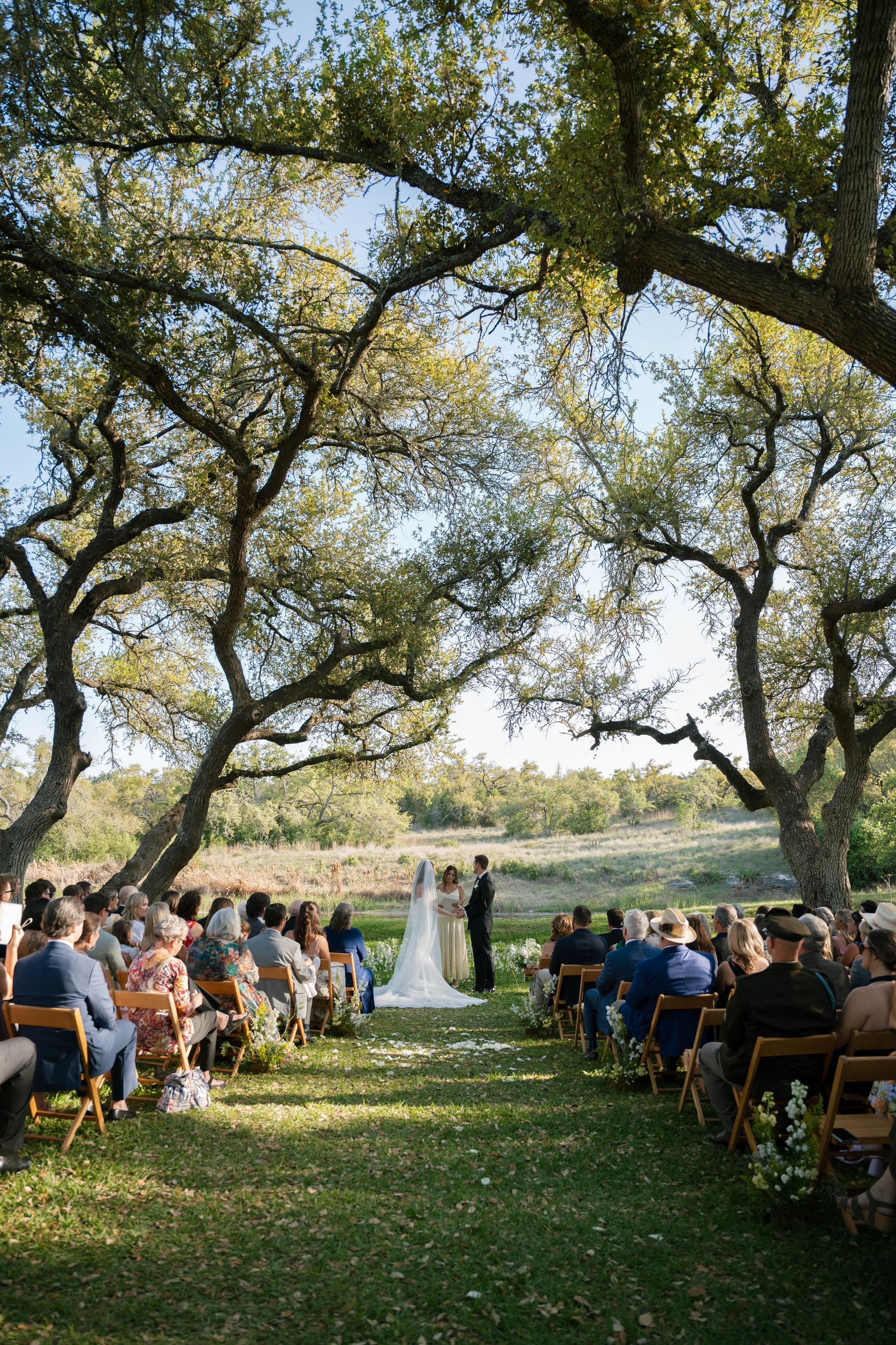 An outdoor wedding ceremony under a canopy of large trees with guests seated in rows facing the couple.