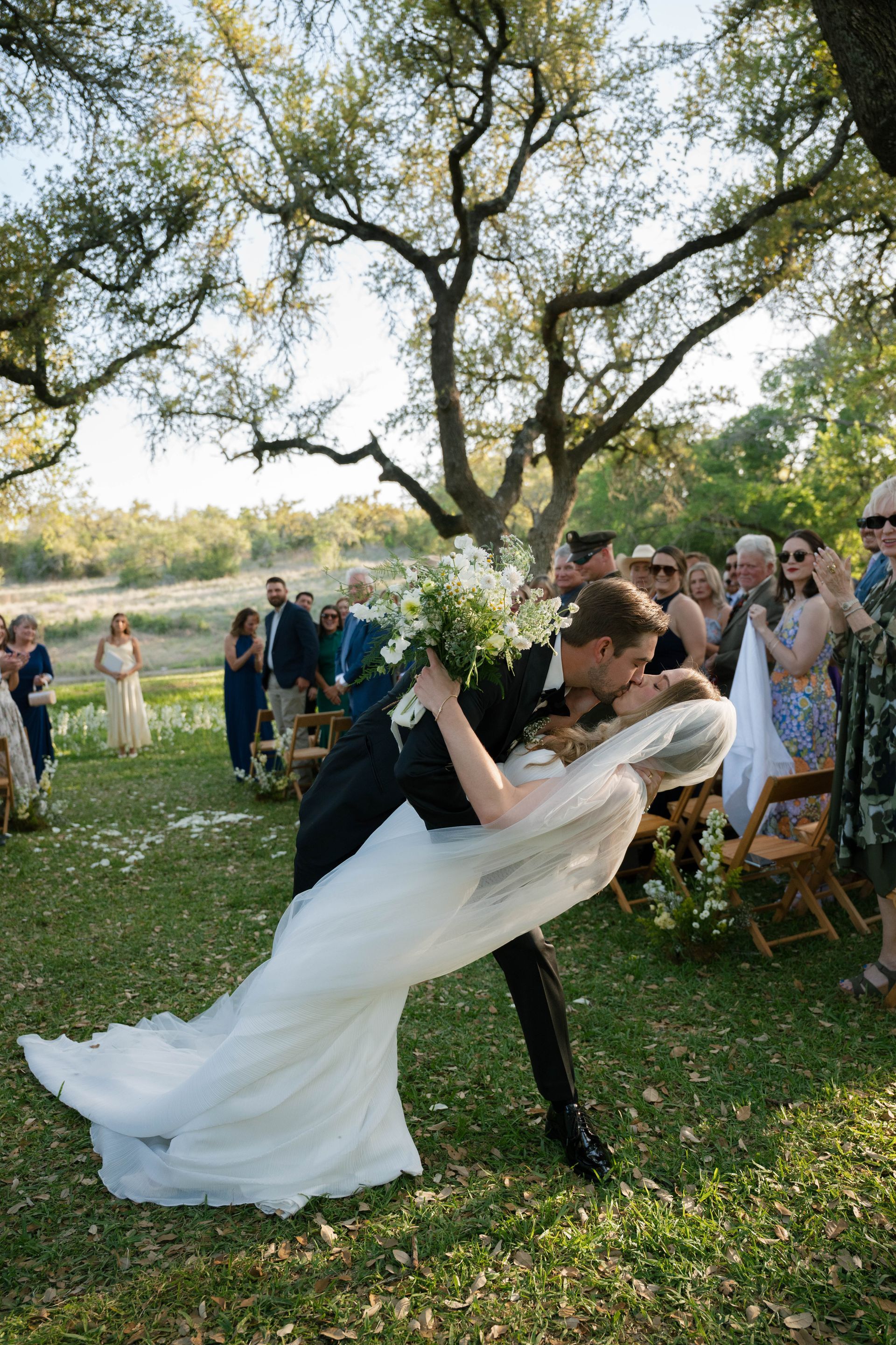 Bride and groom kissing at an outdoor wedding under a large tree, with guests watching nearby.