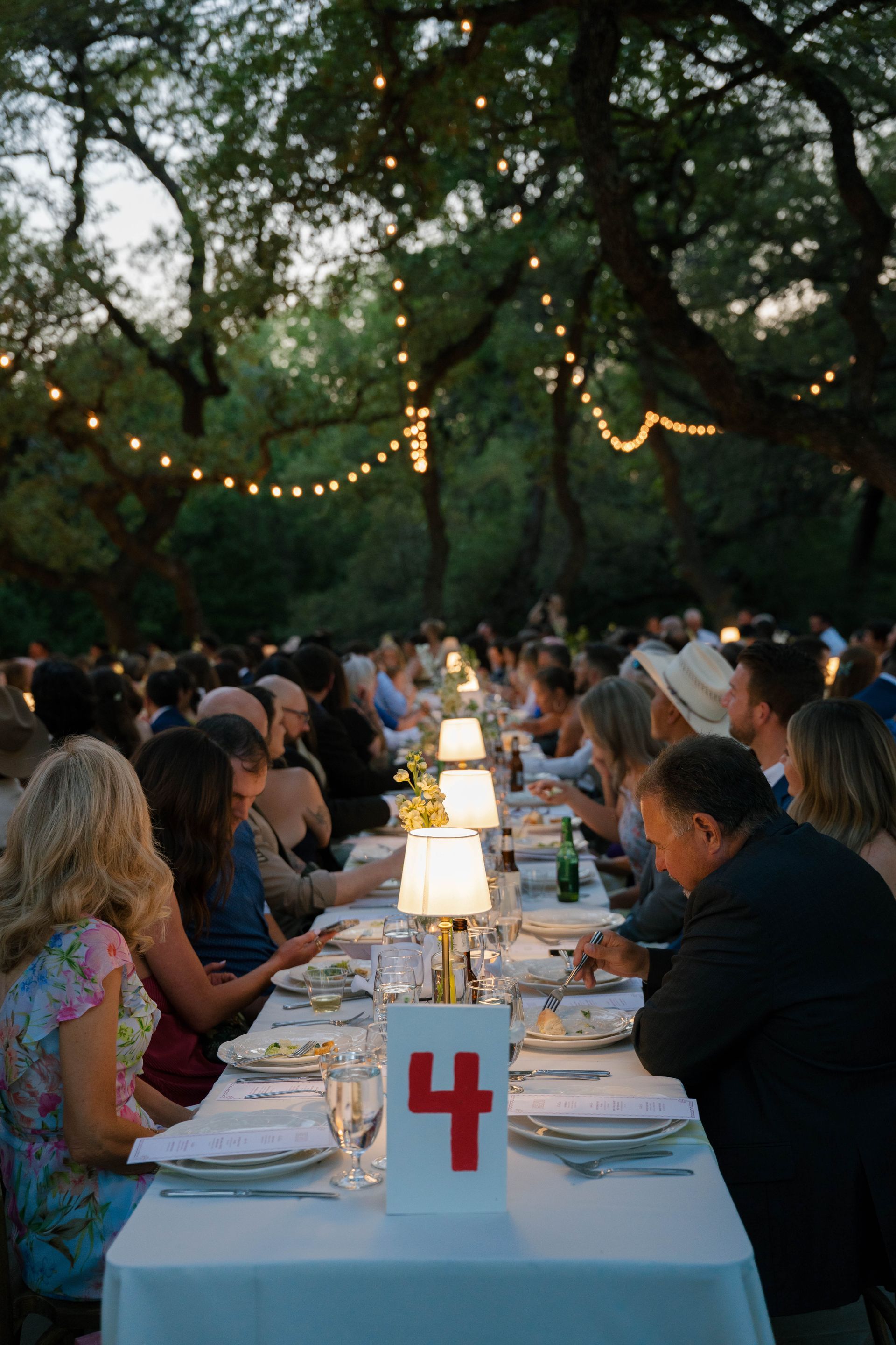 People dine at a long, outdoor table decorated with a red number four card and glowing lamps under string lights.