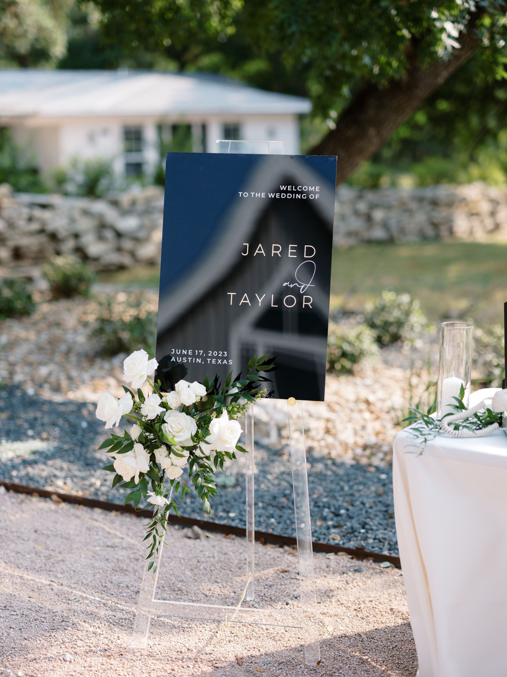 A wedding welcome sign for Sarah and Taylor stands on a clear acrylic easel with white flowers on a gravel path.