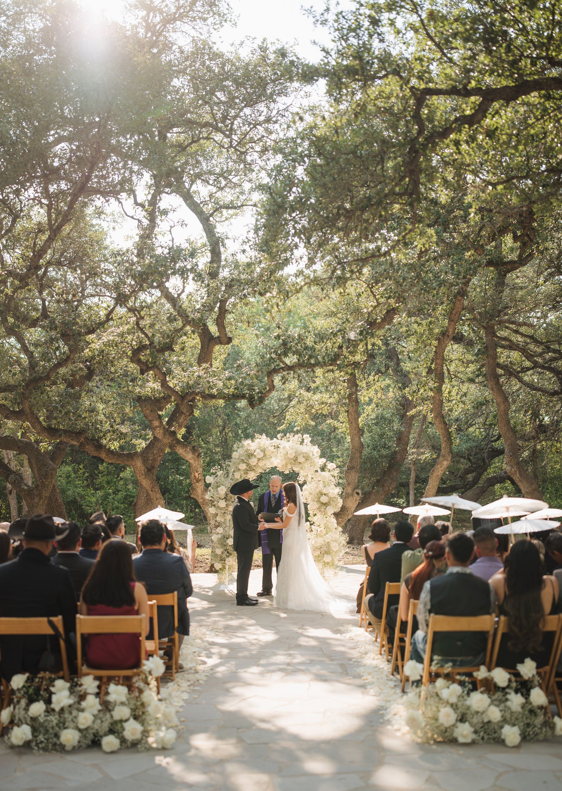 A couple exchanges vows at an outdoor ceremony under an arched floral arrangement surrounded by trees and wedding guests.