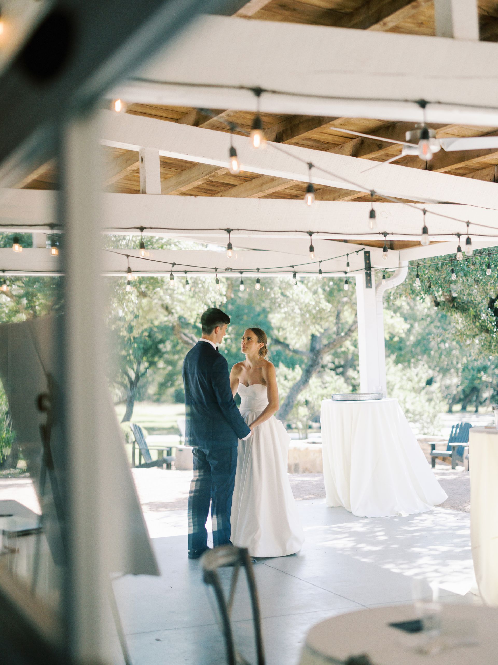 A bride and groom stand facing each other under a covered patio illuminated by hanging string lights.