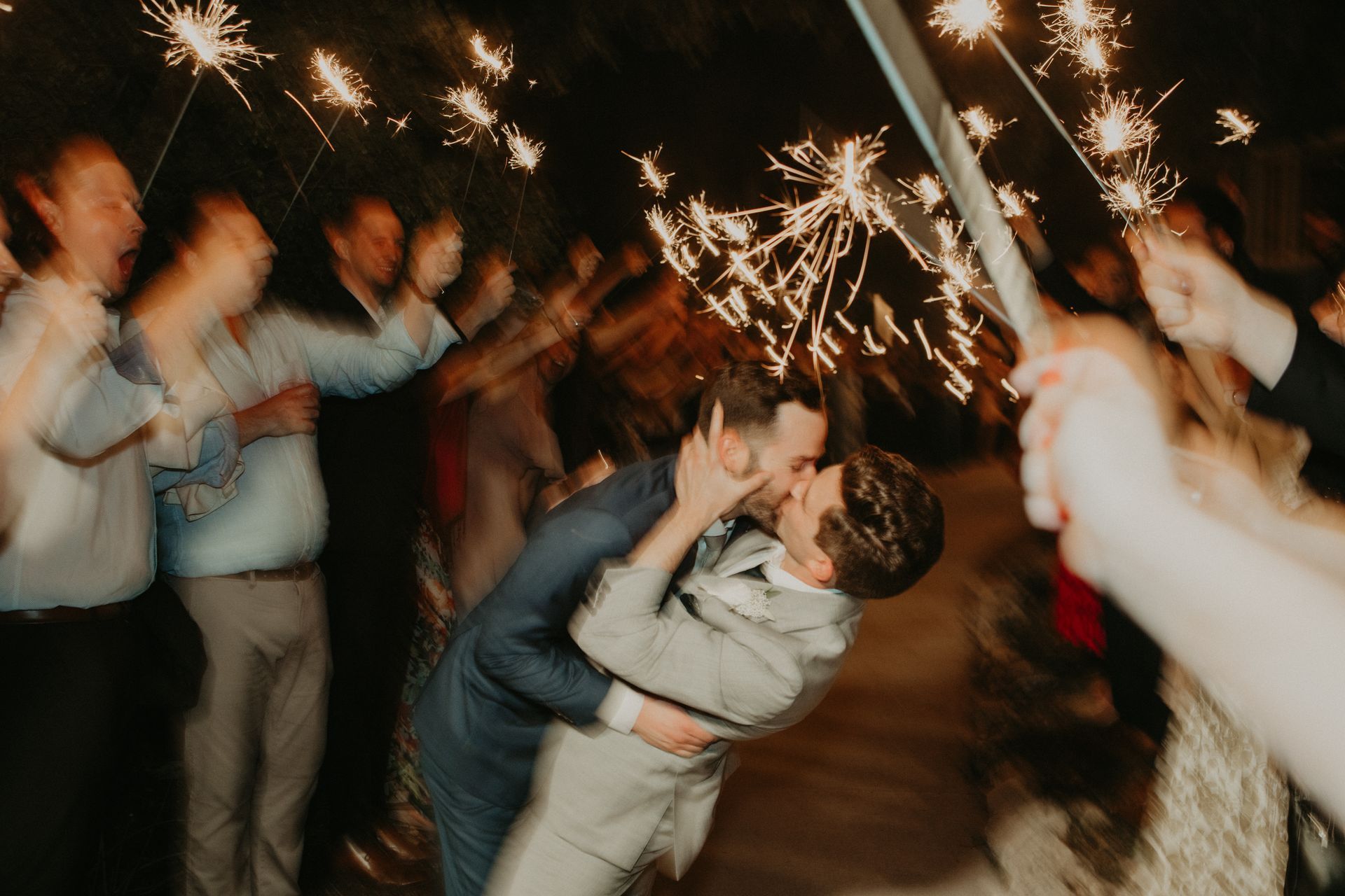 Two people kissing while leaning back, surrounded by people holding lit sparklers at night.