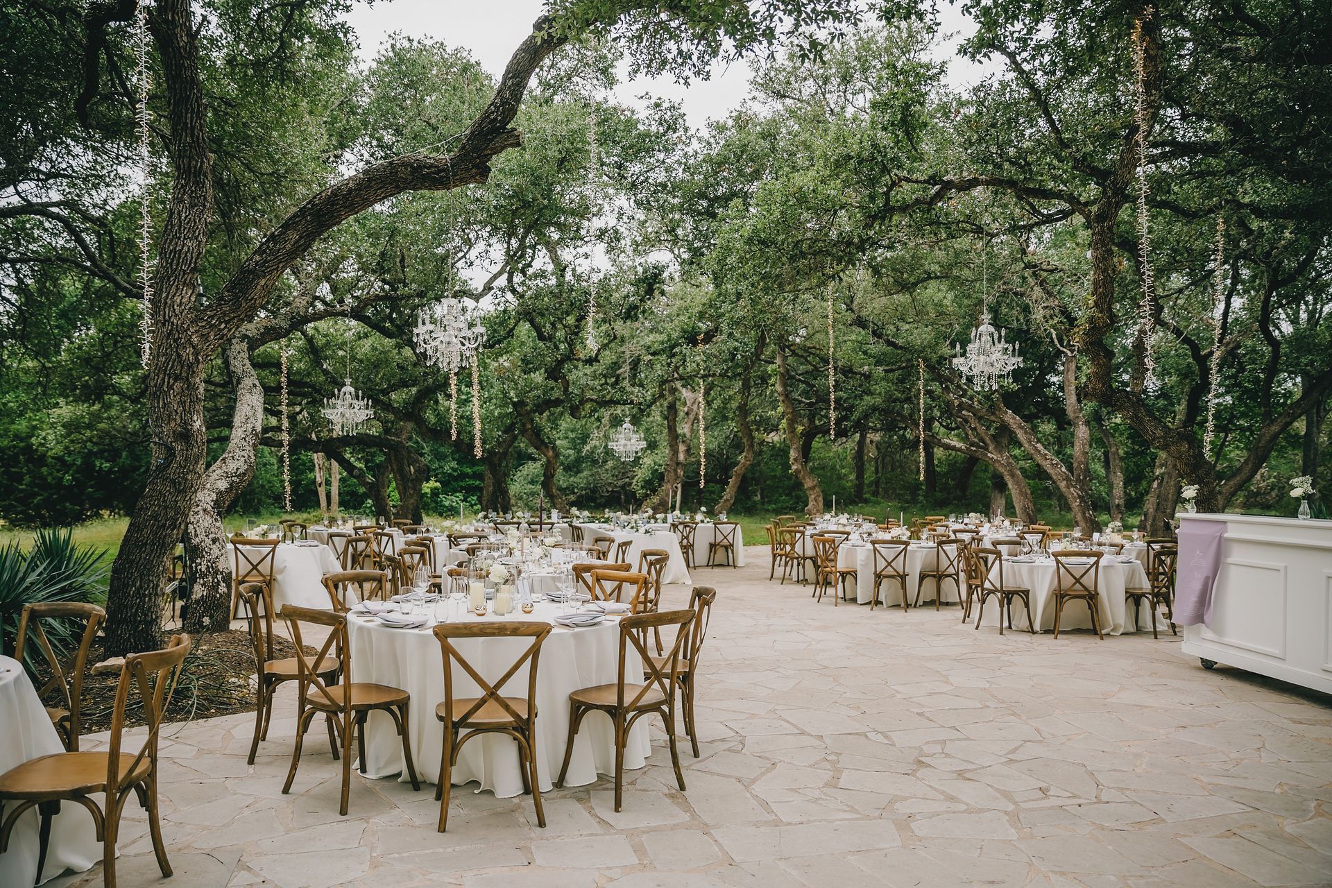 Outdoor wedding reception with white-clothed tables, wooden chairs, and crystal chandeliers hanging from oak trees.
