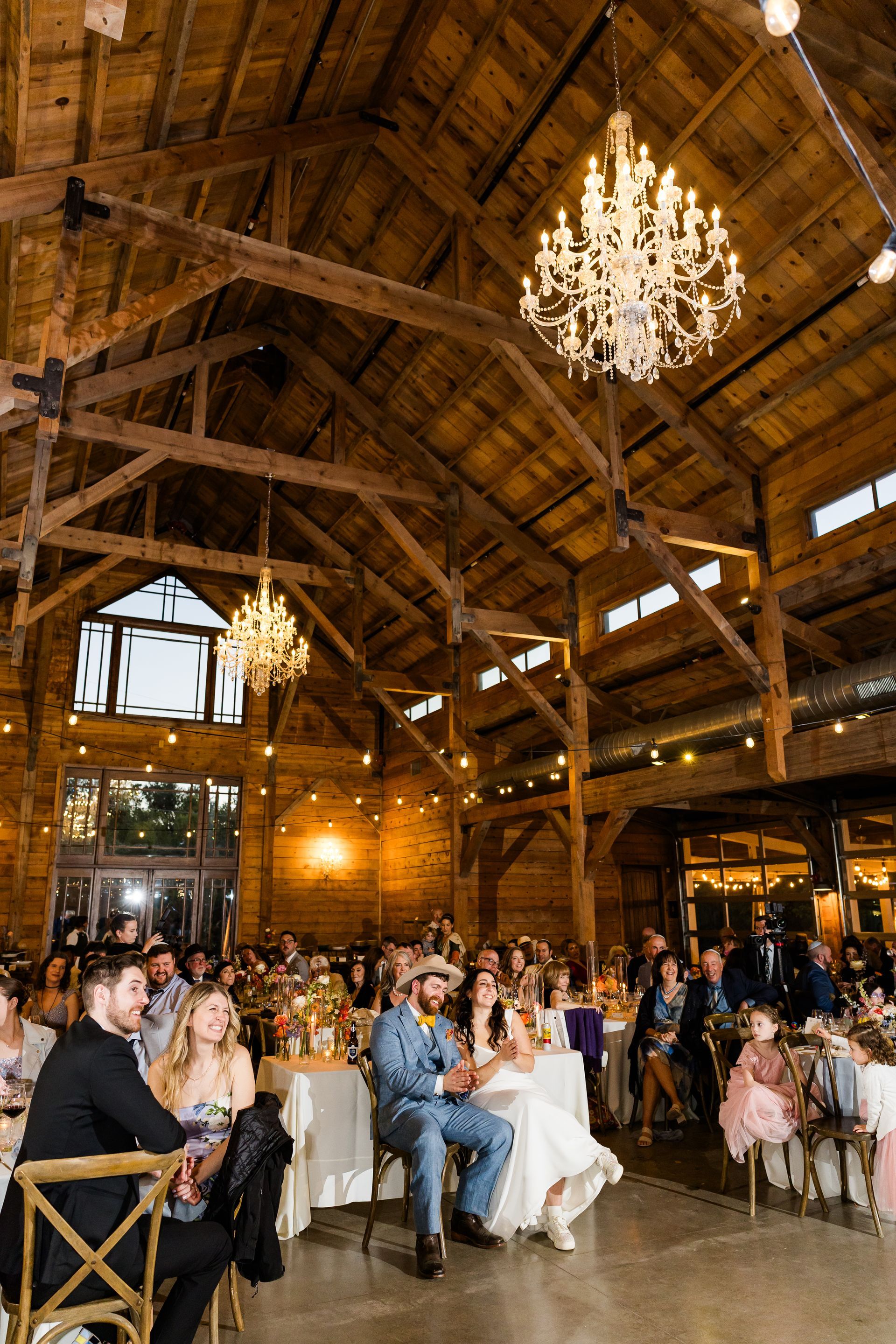 A rustic wooden barn wedding reception features guests seated at round tables under glowing chandeliers and timber beams.