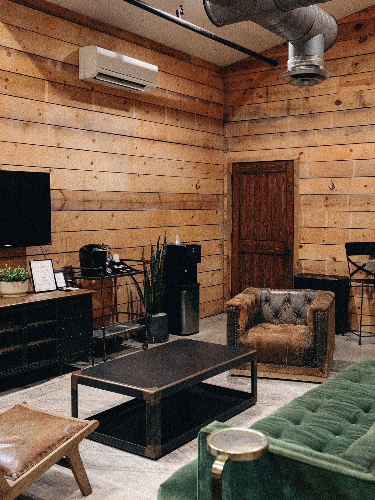 A rustic lounge with log walls, a green tufted sofa, brown leather armchair, coffee table, and exposed ceiling ductwork.
