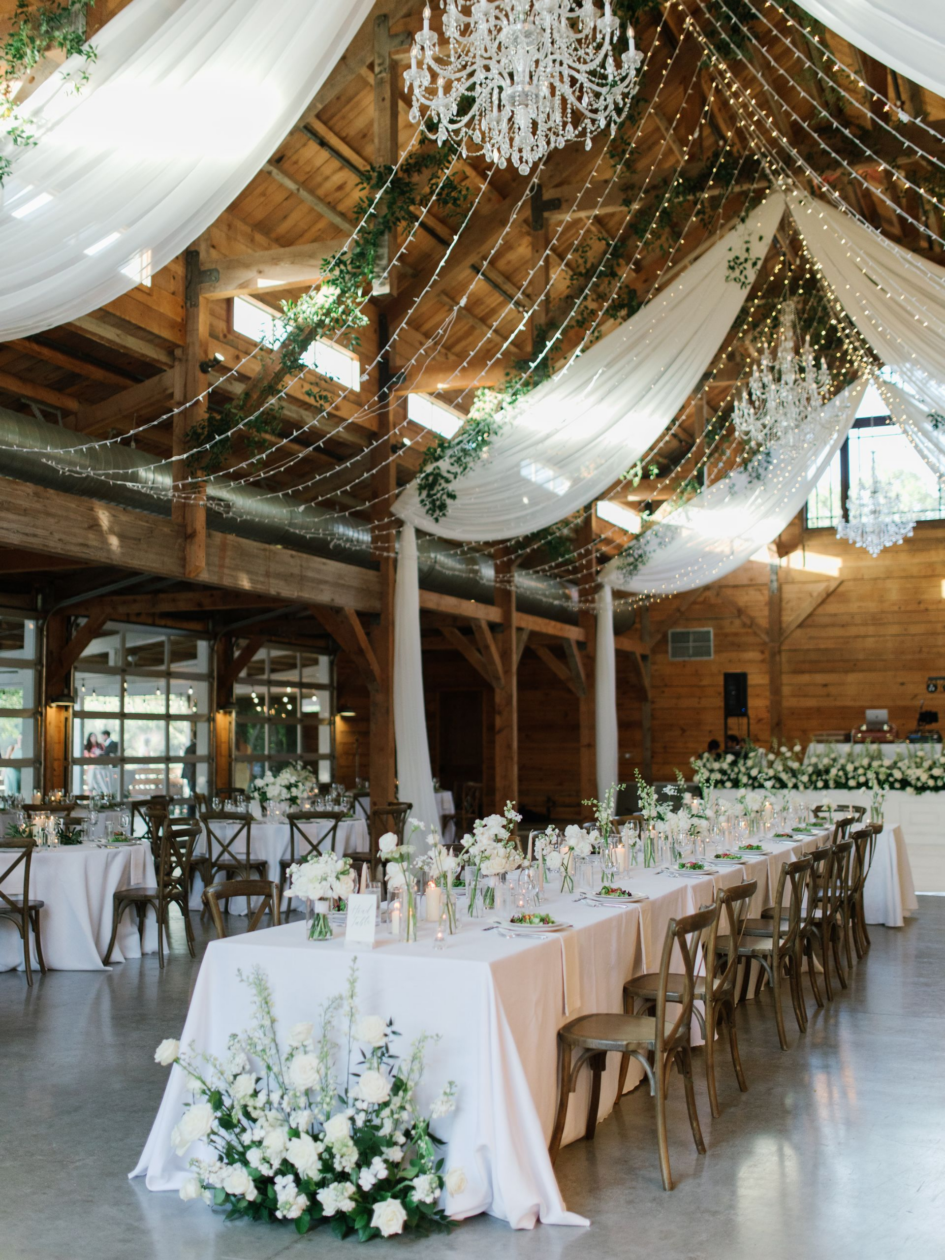 A long wedding table with white linens and floral arrangements in a rustic wooden barn with draped fabric and chandeliers.