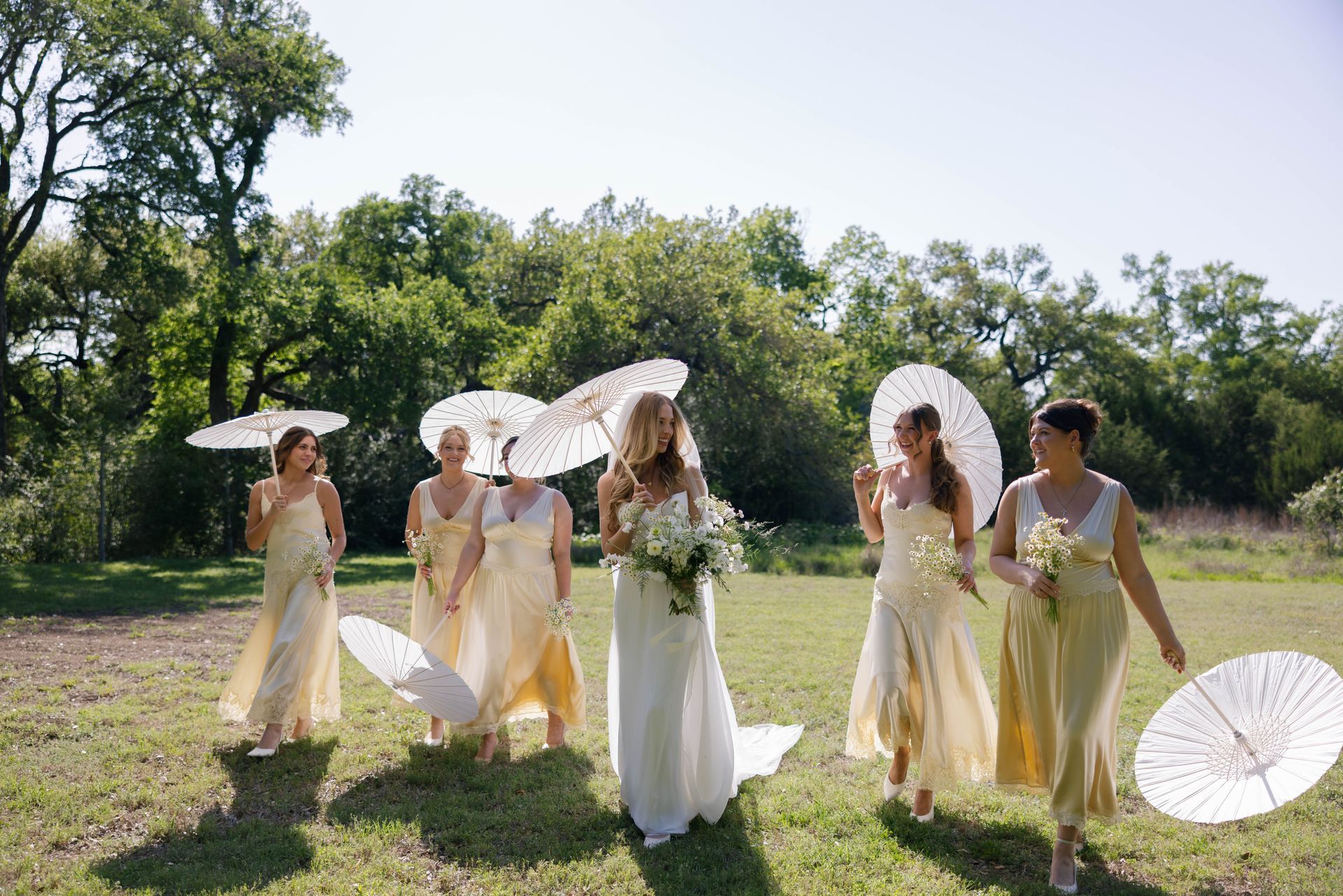 A bride and four bridesmaids in matching yellow dresses walk outdoors, carrying white parasols on a sunny, grassy field.