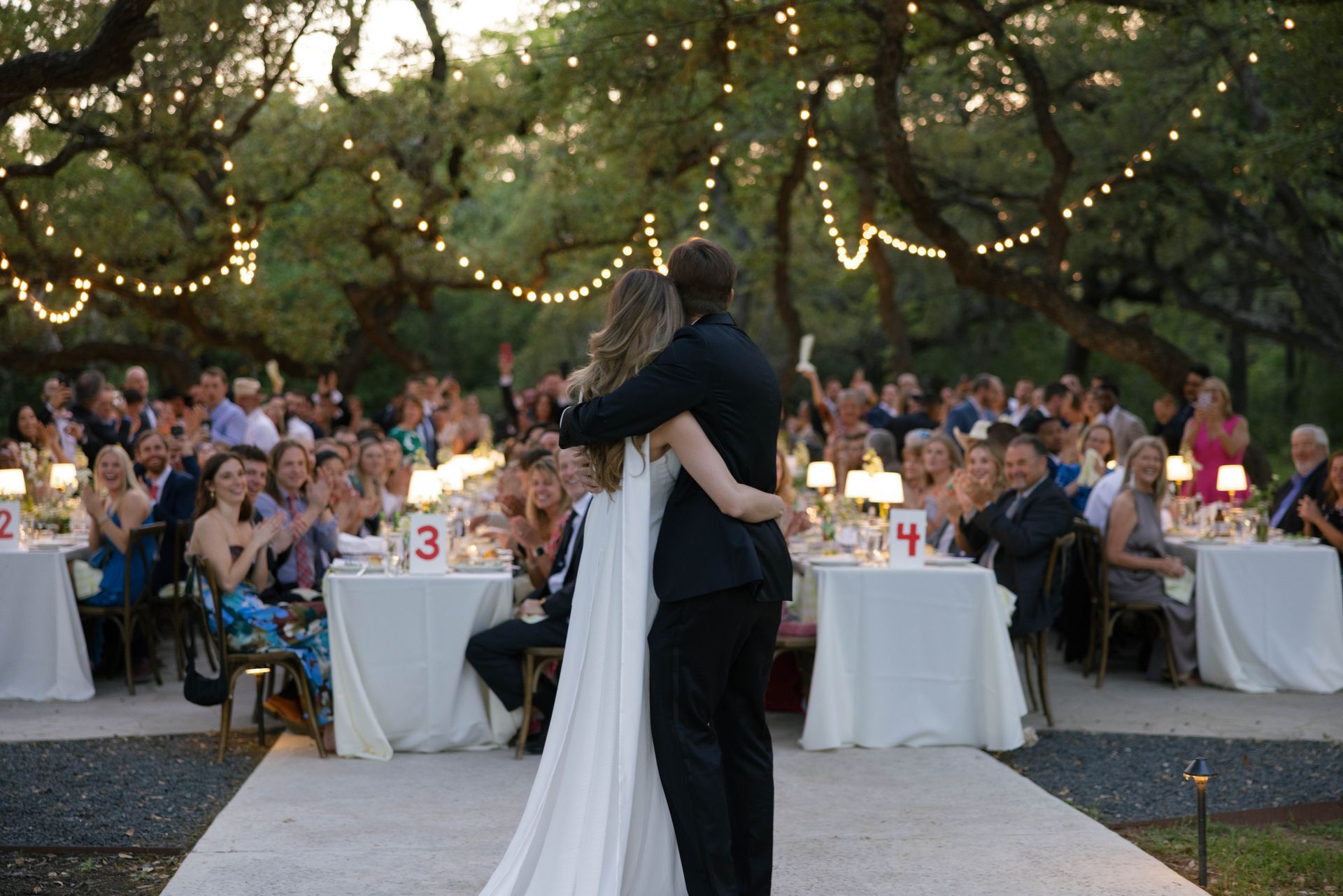 A couple dances at an outdoor wedding reception with guests seated at tables under string lights and trees.