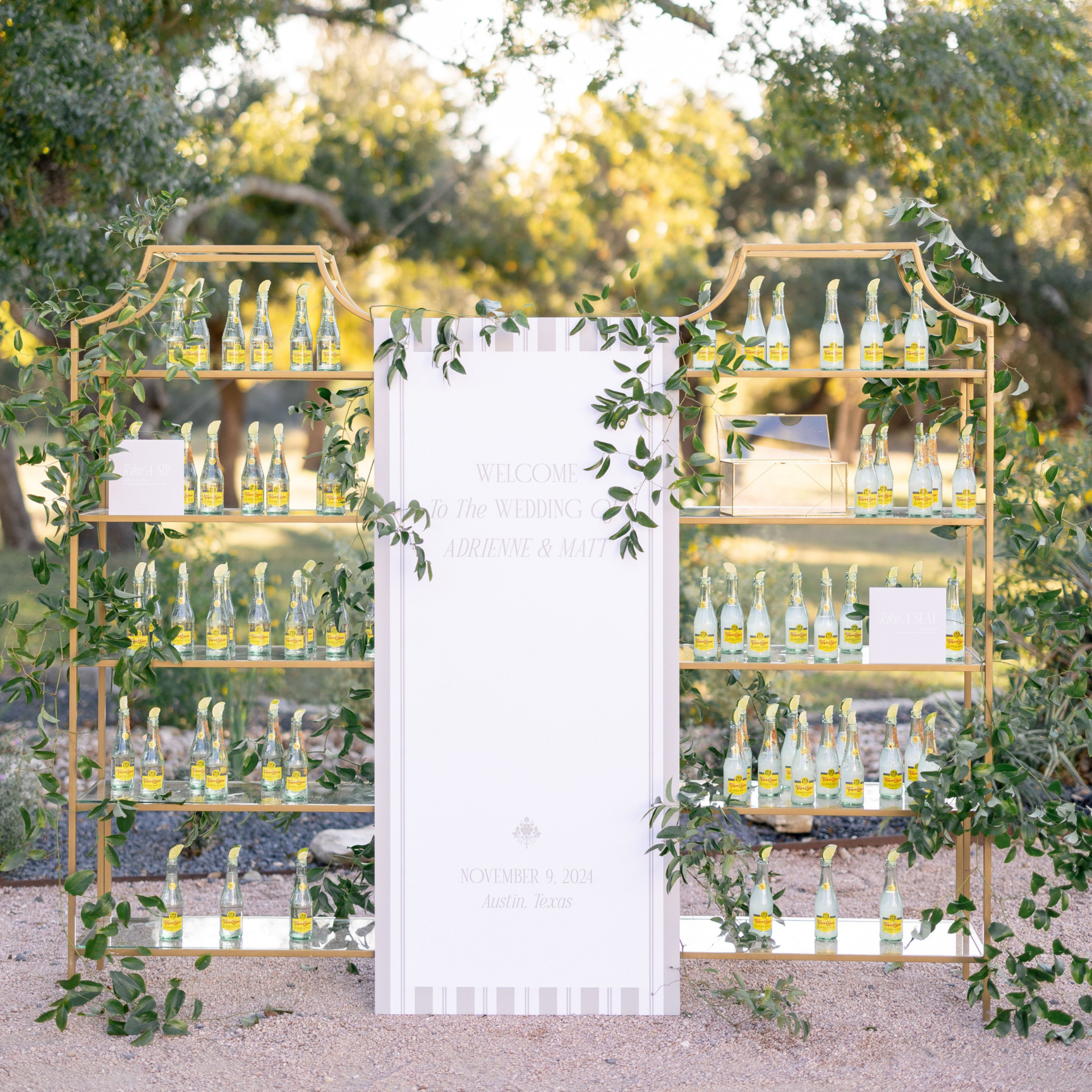 An outdoor gold shelving unit adorned with greenery, holding rows of glass bottles, flanking a central event sign.