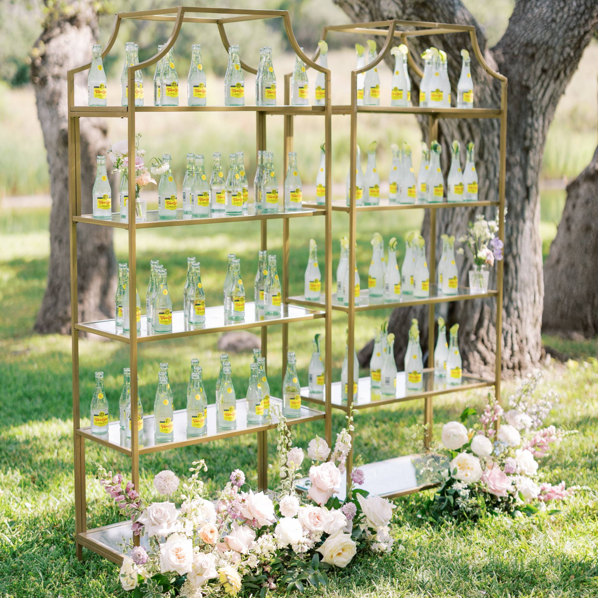 Gold shelving units stand outdoors, filled with rows of bottled drinks, decorated with lush bouquets of pale flowers.