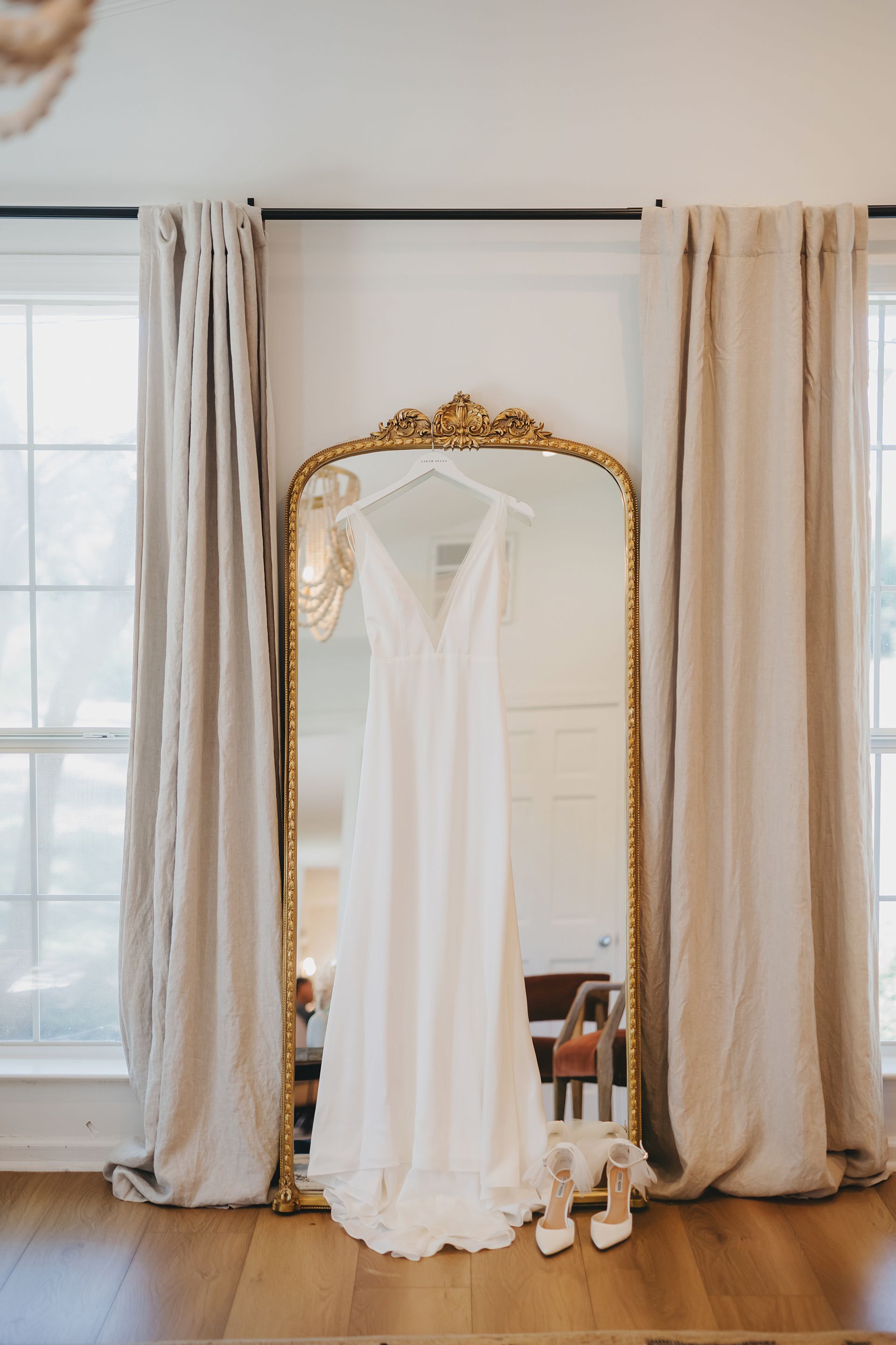 A white wedding dress hangs from a gold-framed mirror between two beige curtains, with bridal heels placed at the base.