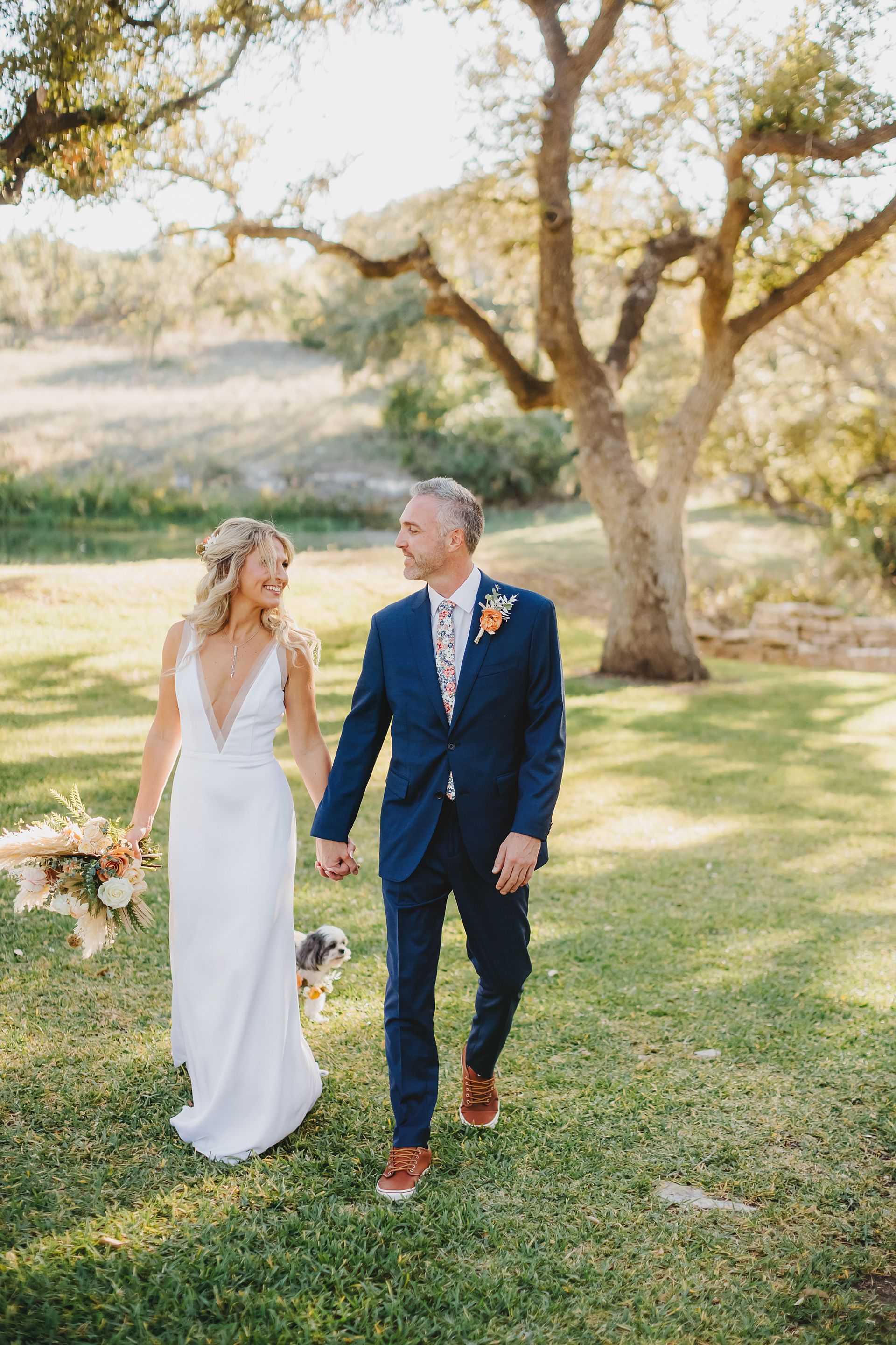 A bride and groom walk hand-in-hand through a grassy field under a large tree, carrying a bouquet with a small dog nearby.