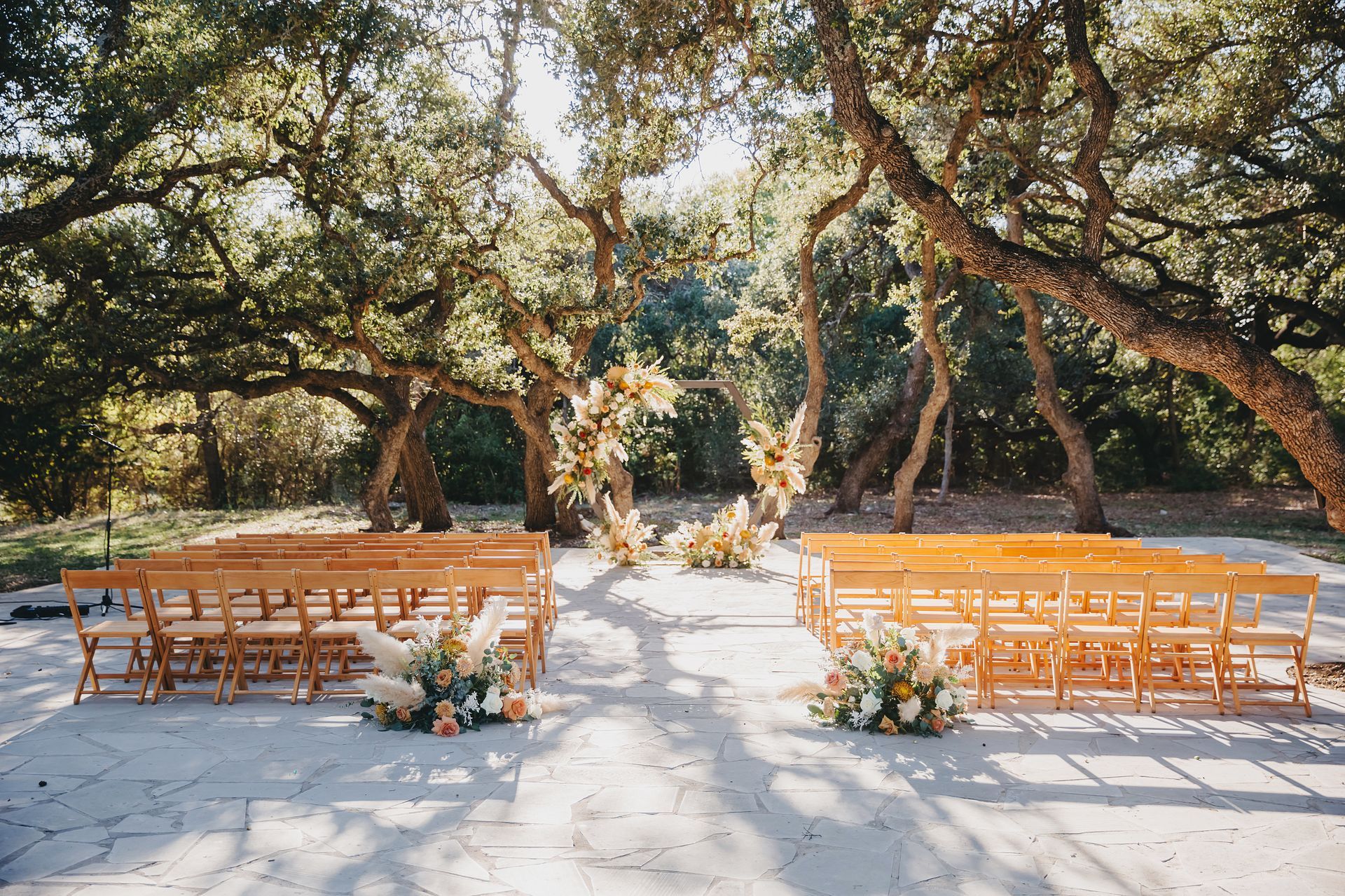 Outdoor wedding ceremony setup with wooden chairs arranged in rows facing a circular floral arch under mature oak trees.