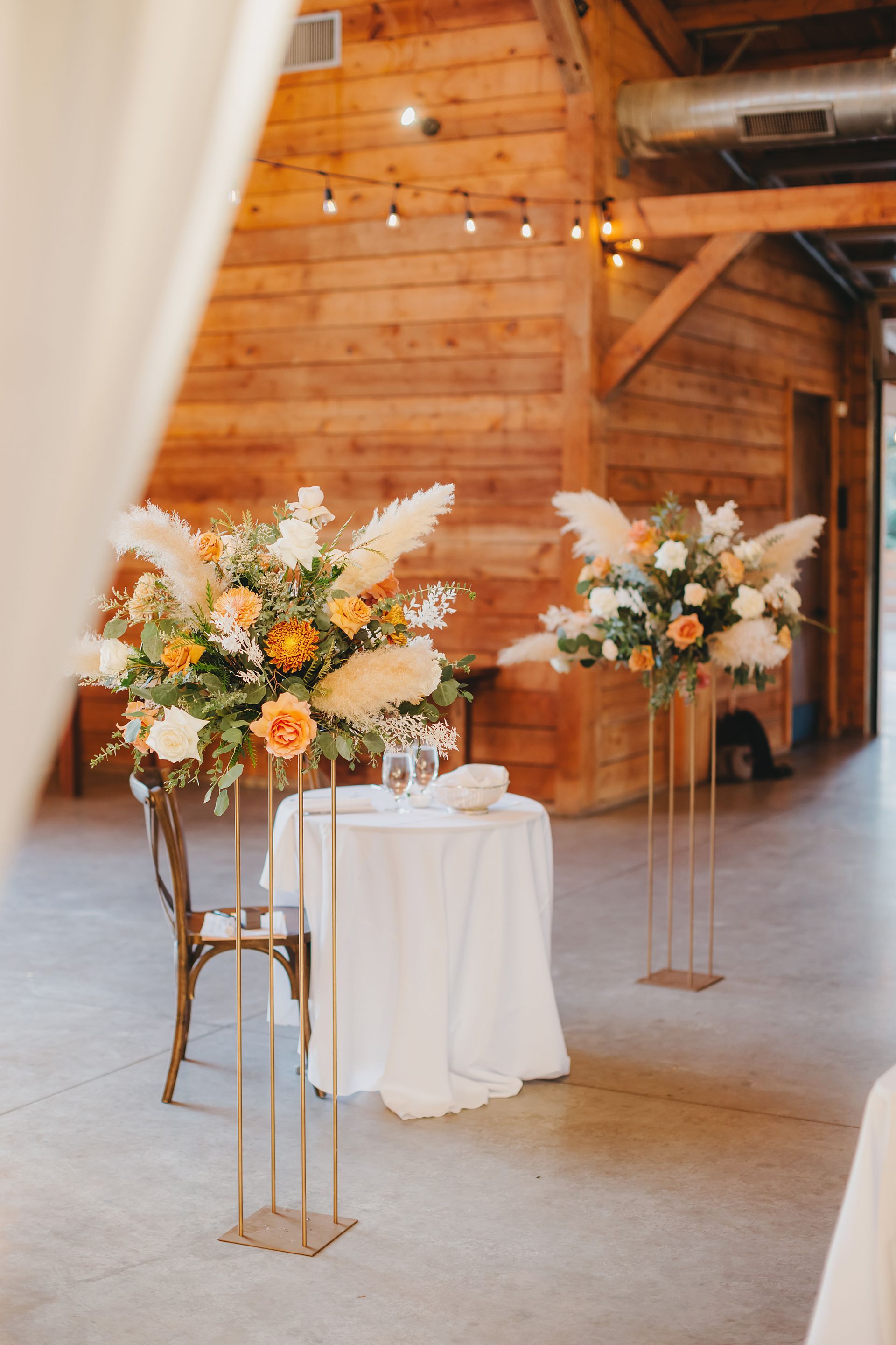 Two floral arrangements featuring pampas grass and orange roses on tall gold stands next to a small white-covered table at The Addison Grove.