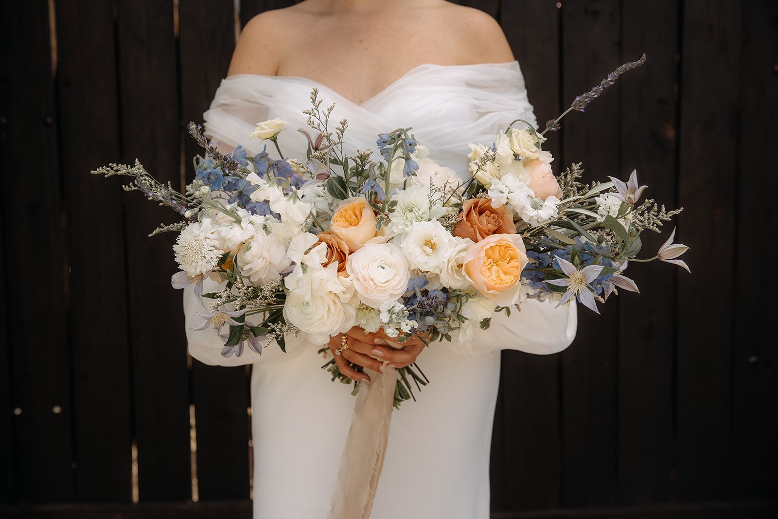 A bride in a white off-the-shoulder dress holds a large, asymmetrical bouquet of white, peach, and dusty blue flowers.
