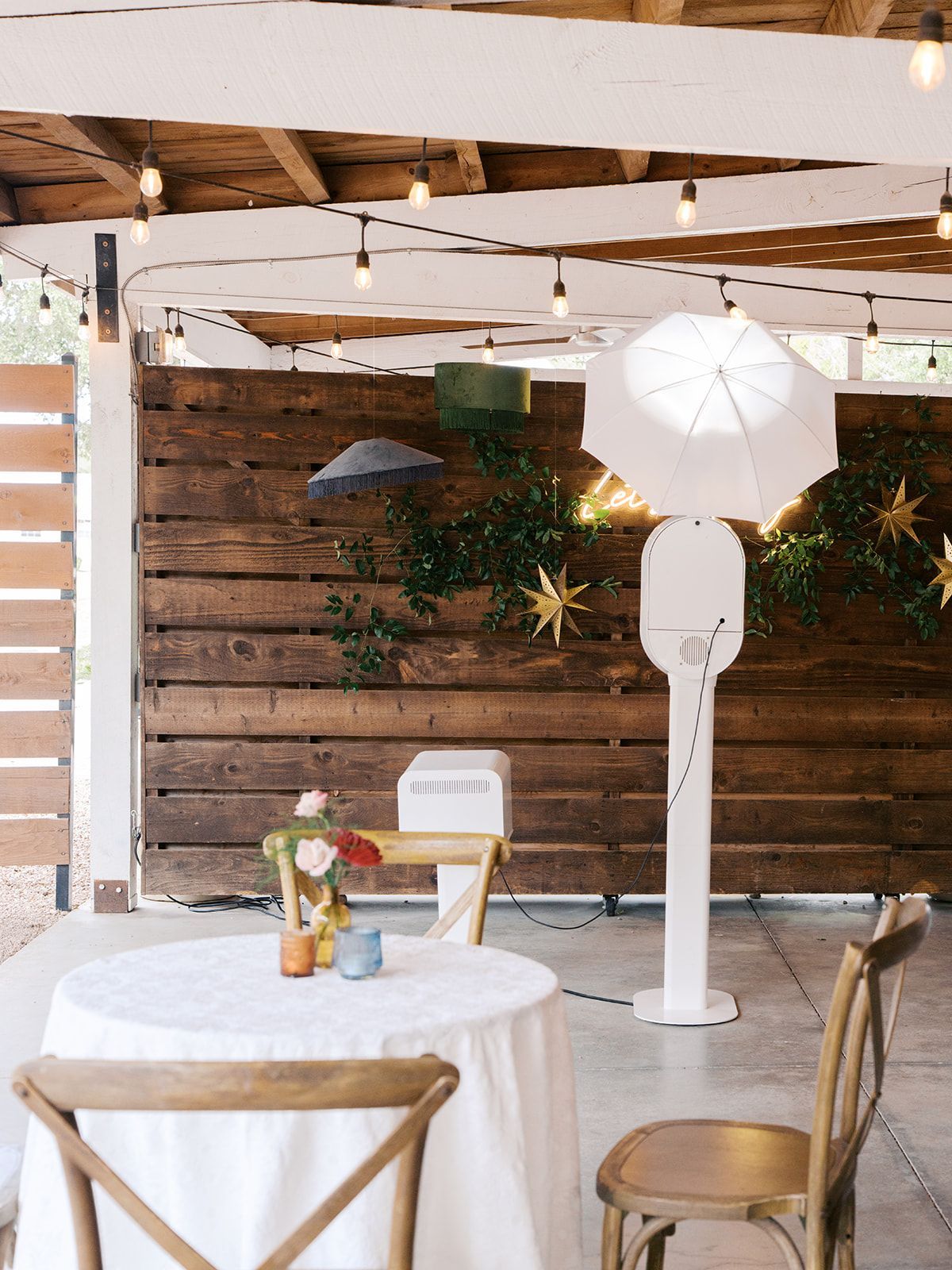 A photo booth setup with a white umbrella light stands on a patio with wooden walls, bistro lights, and a table with chairs.