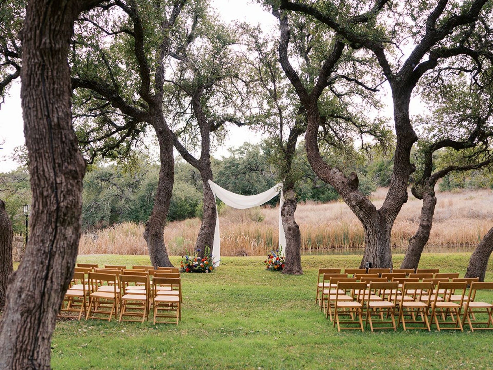 An outdoor wedding ceremony setup with rows of wooden chairs facing a draped white arbor nestled between large trees.