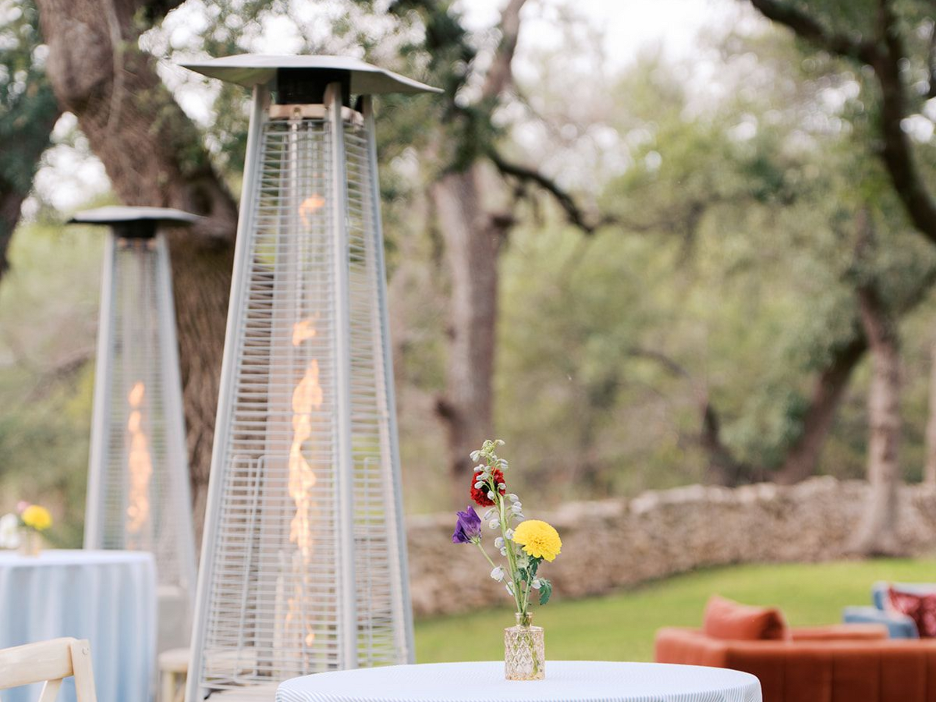 Outdoor event space with tall, pyramid-shaped heaters, bistro tables with small floral centerpieces, and lawn furniture.
