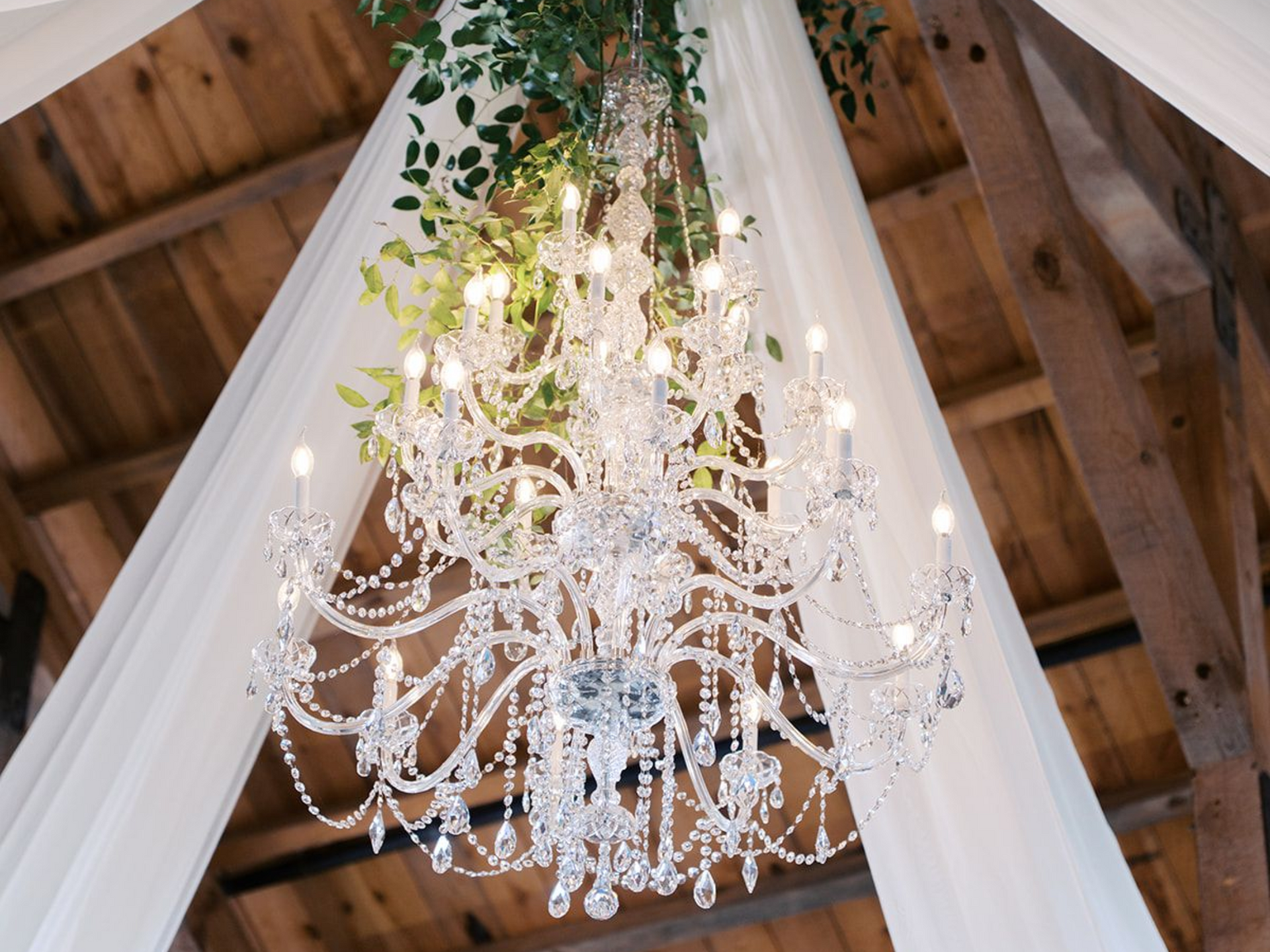 An ornate crystal chandelier hangs from a rustic wooden ceiling, draped with white fabric and decorative greenery.