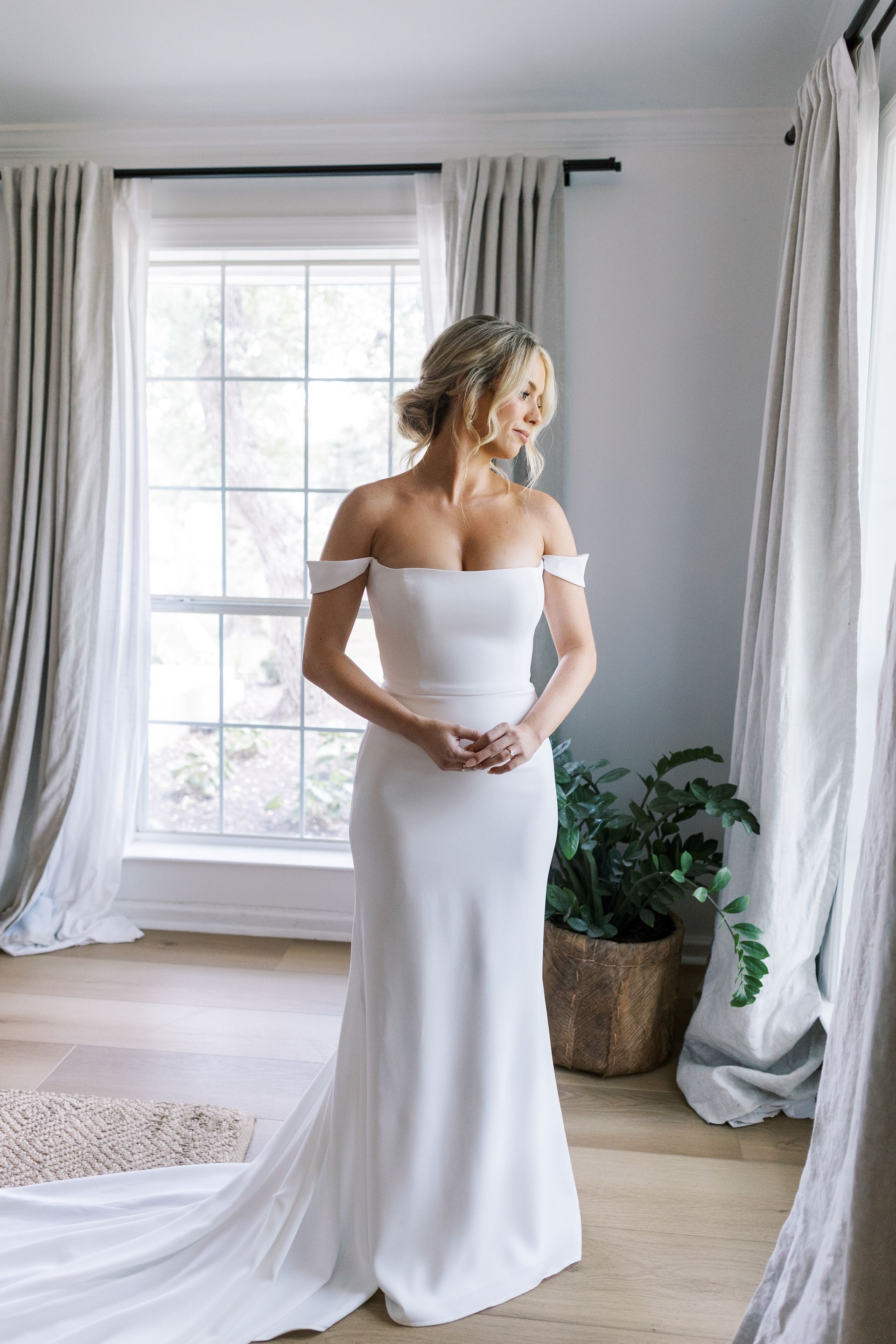 A person in a white, off-the-shoulder wedding dress stands by a window in a bright room with curtains and a potted plant.