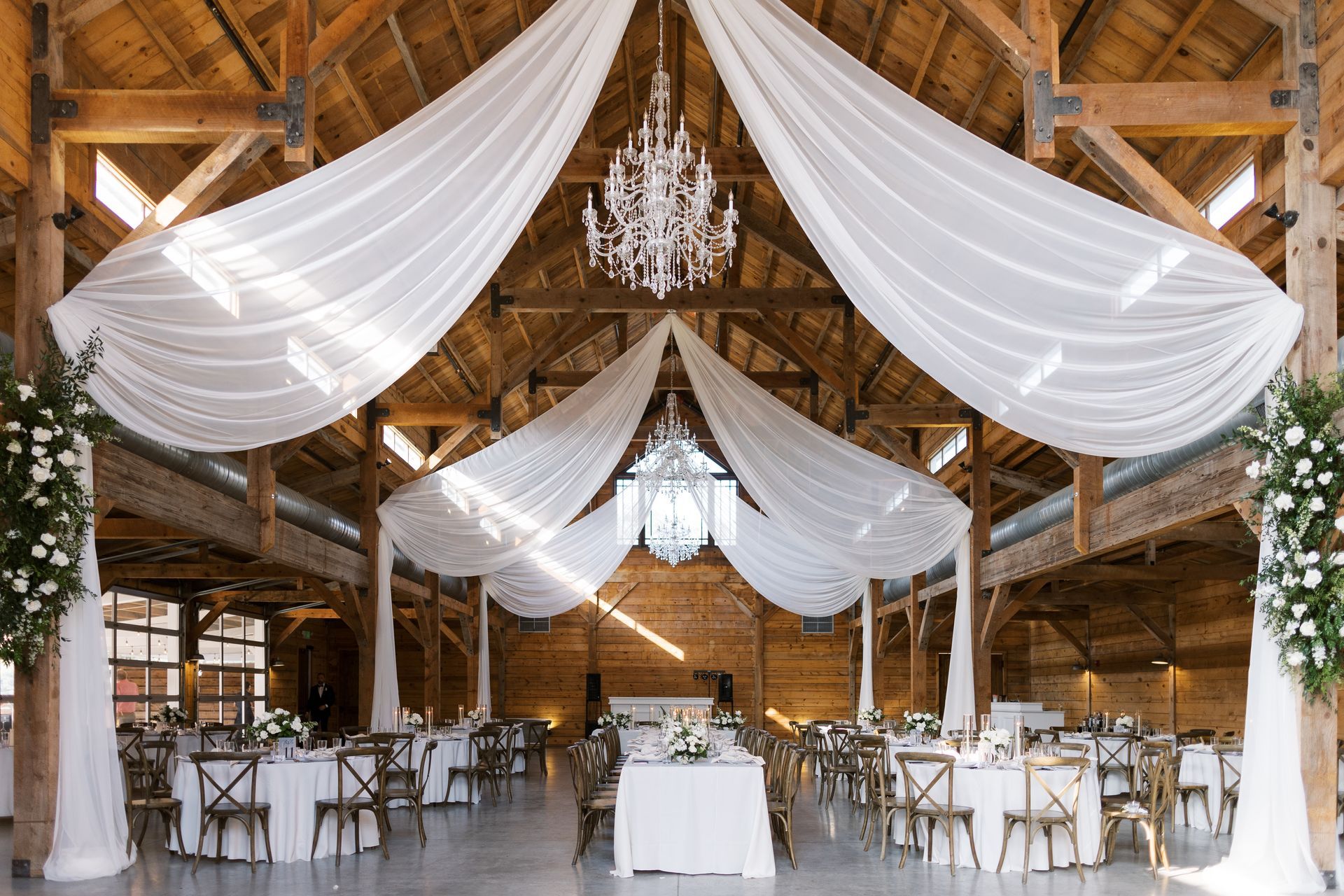 A rustic, timber-framed barn venue set for a wedding with white draped fabric, crystal chandeliers, and round tables.