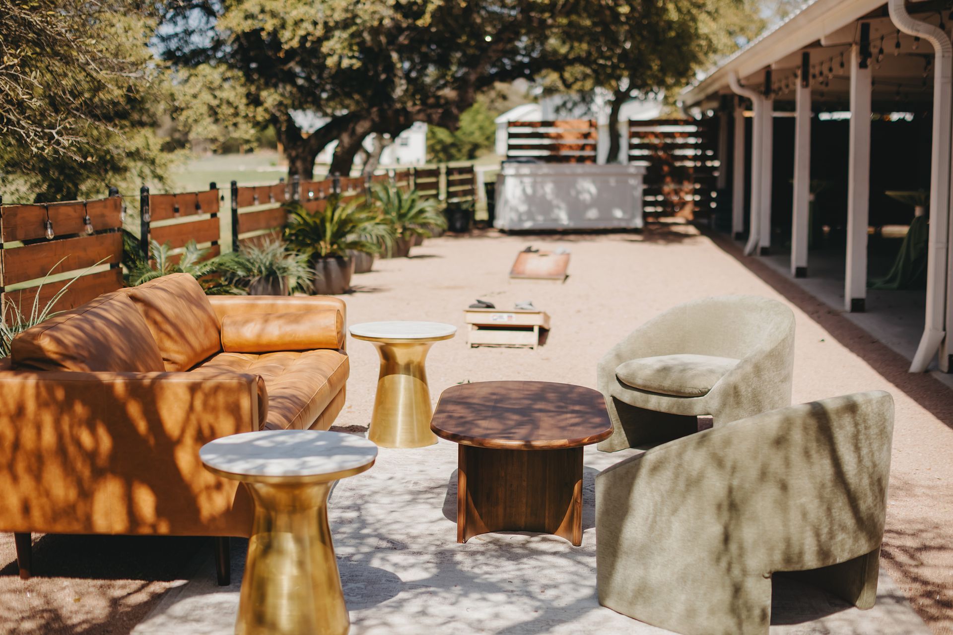 An outdoor lounge area featuring a brown leather sofa, olive chairs, brass side tables, and a coffee table on a patio.