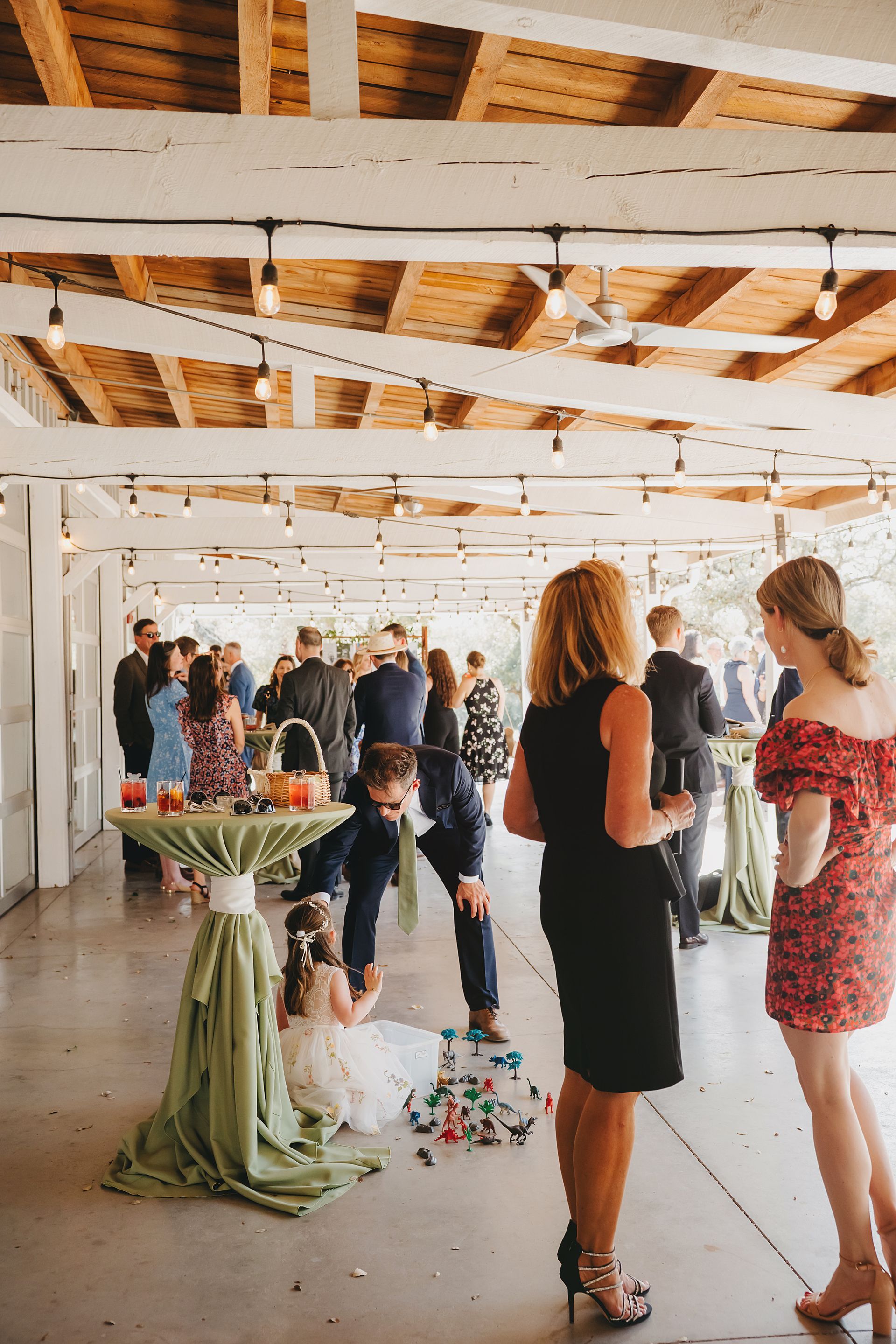 People socialize at an outdoor reception under a covered patio with string lights, while a child sits near a draped table.