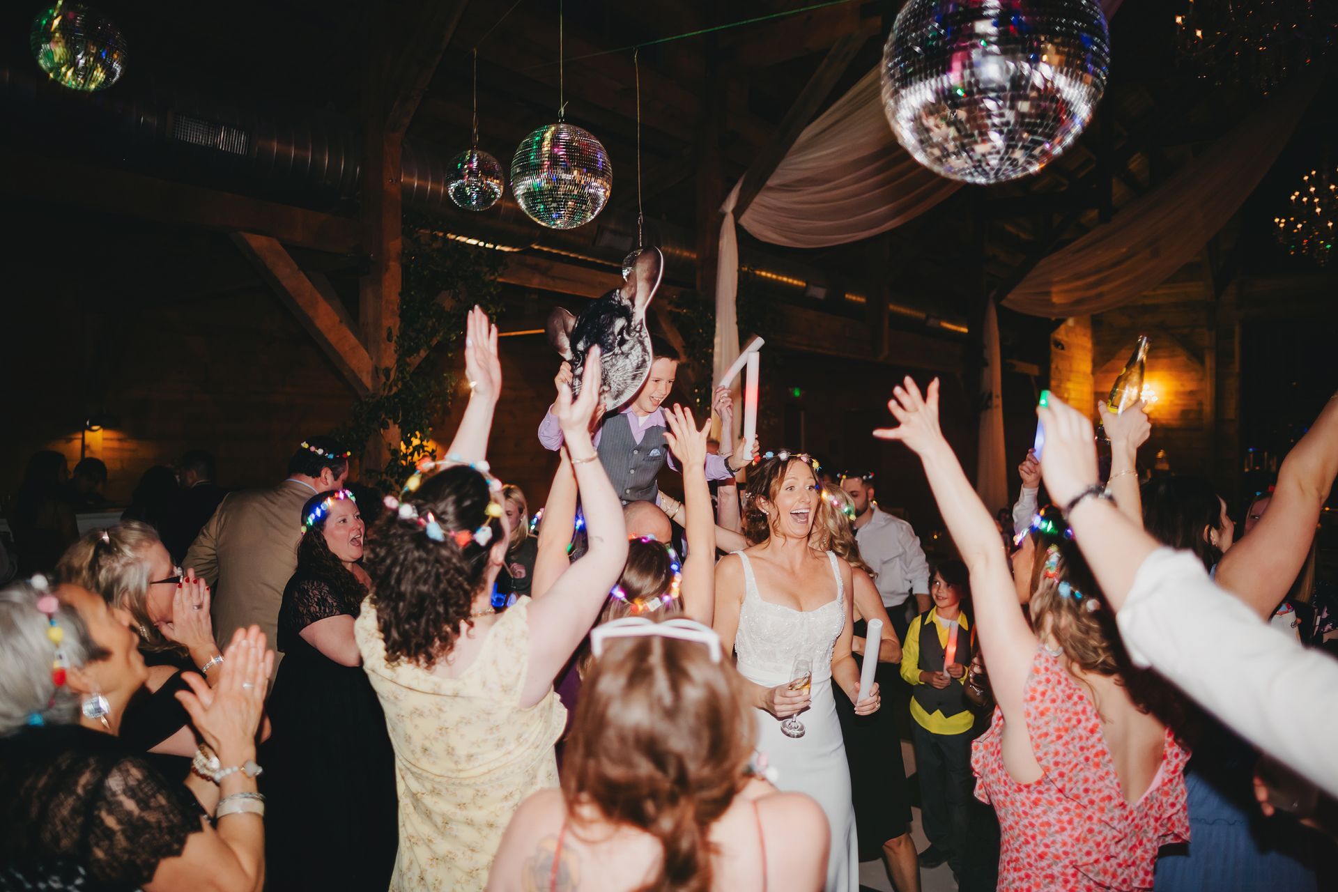 A festive wedding reception with guests dancing under disco balls as a person is lifted into the air.