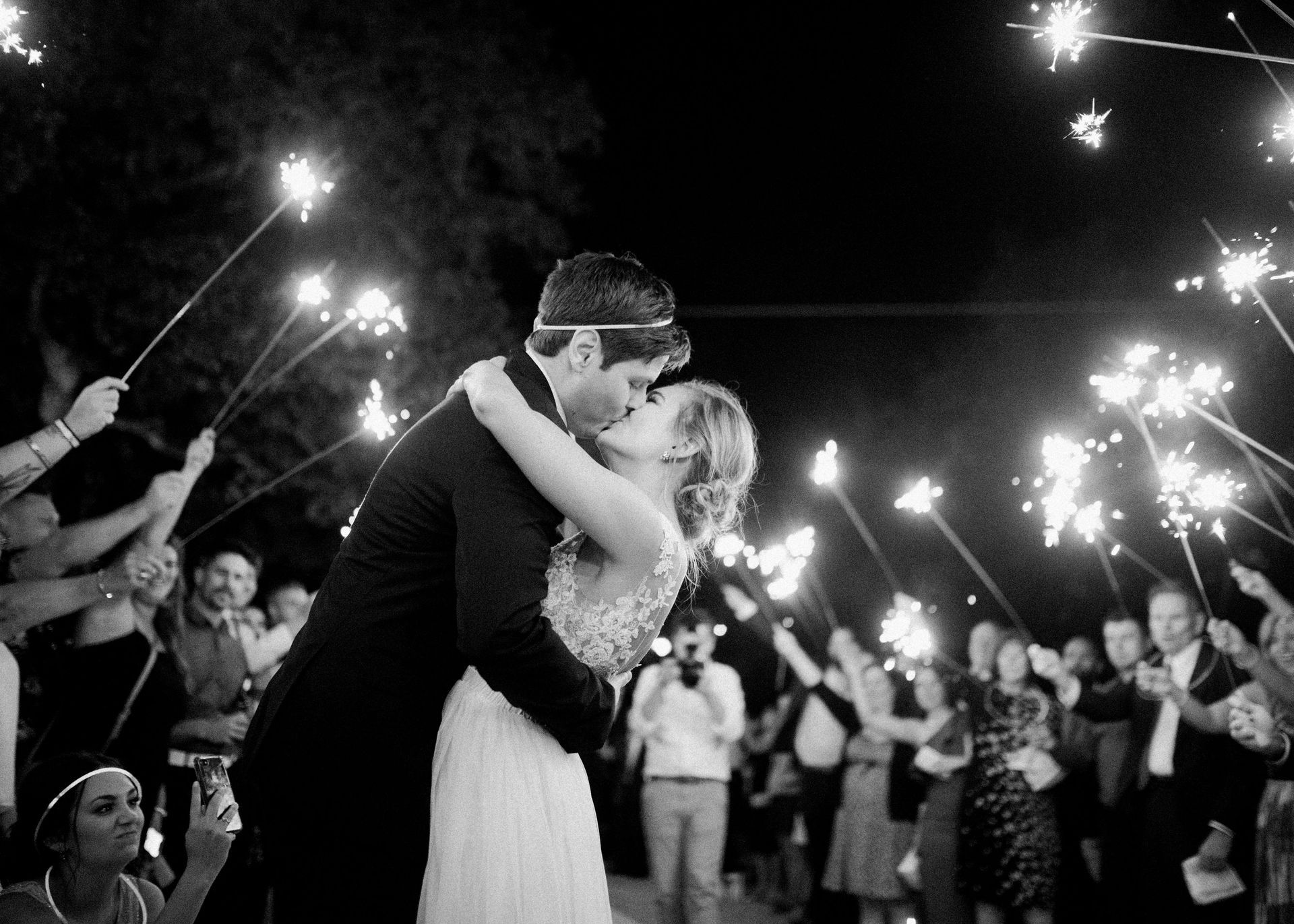 A newlywed couple kisses during a nighttime wedding exit, surrounded by guests holding glowing sparklers.