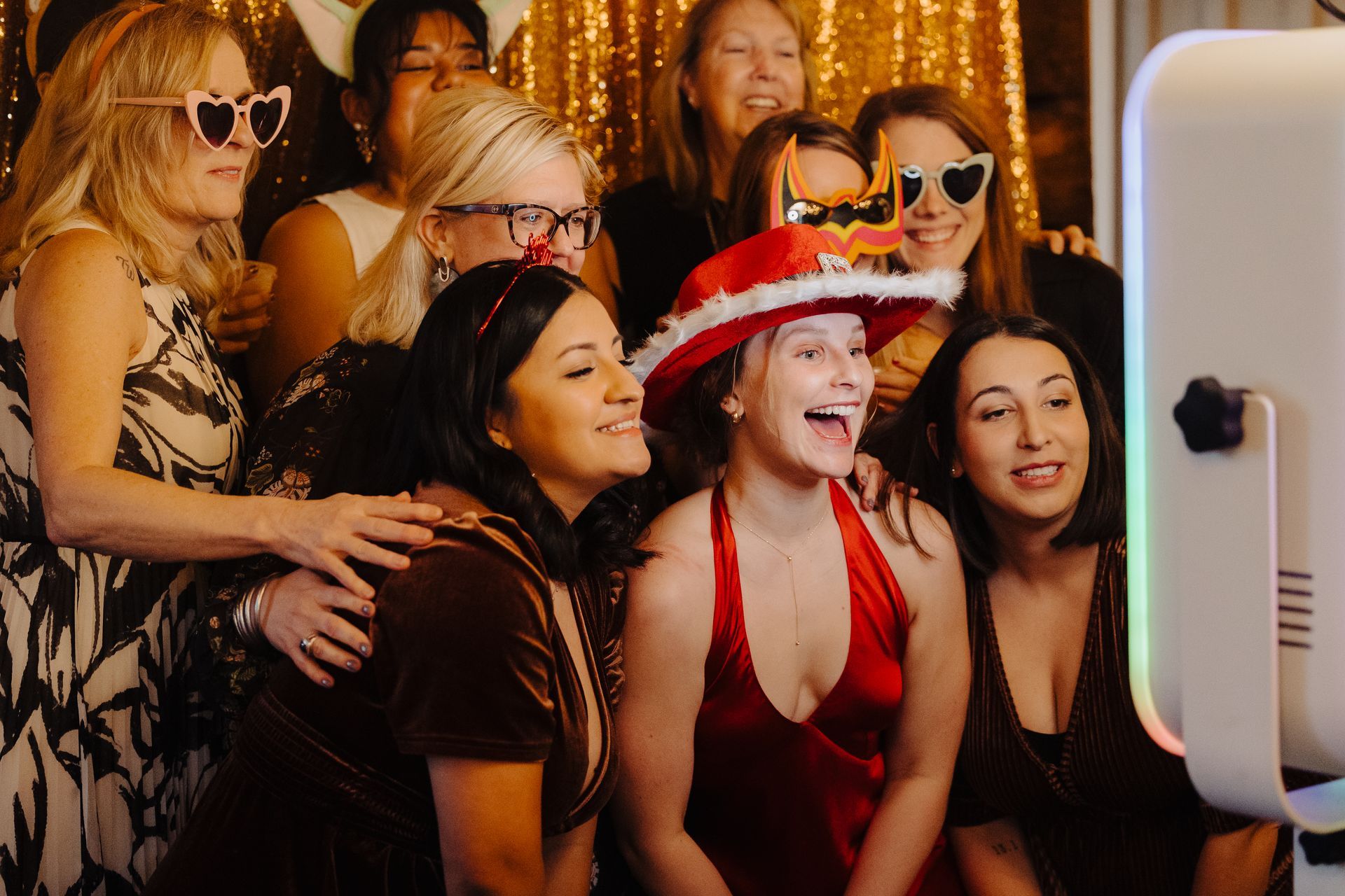 A group of people wearing party props and hats pose for a photo at a photo booth against a gold, sequined backdrop.