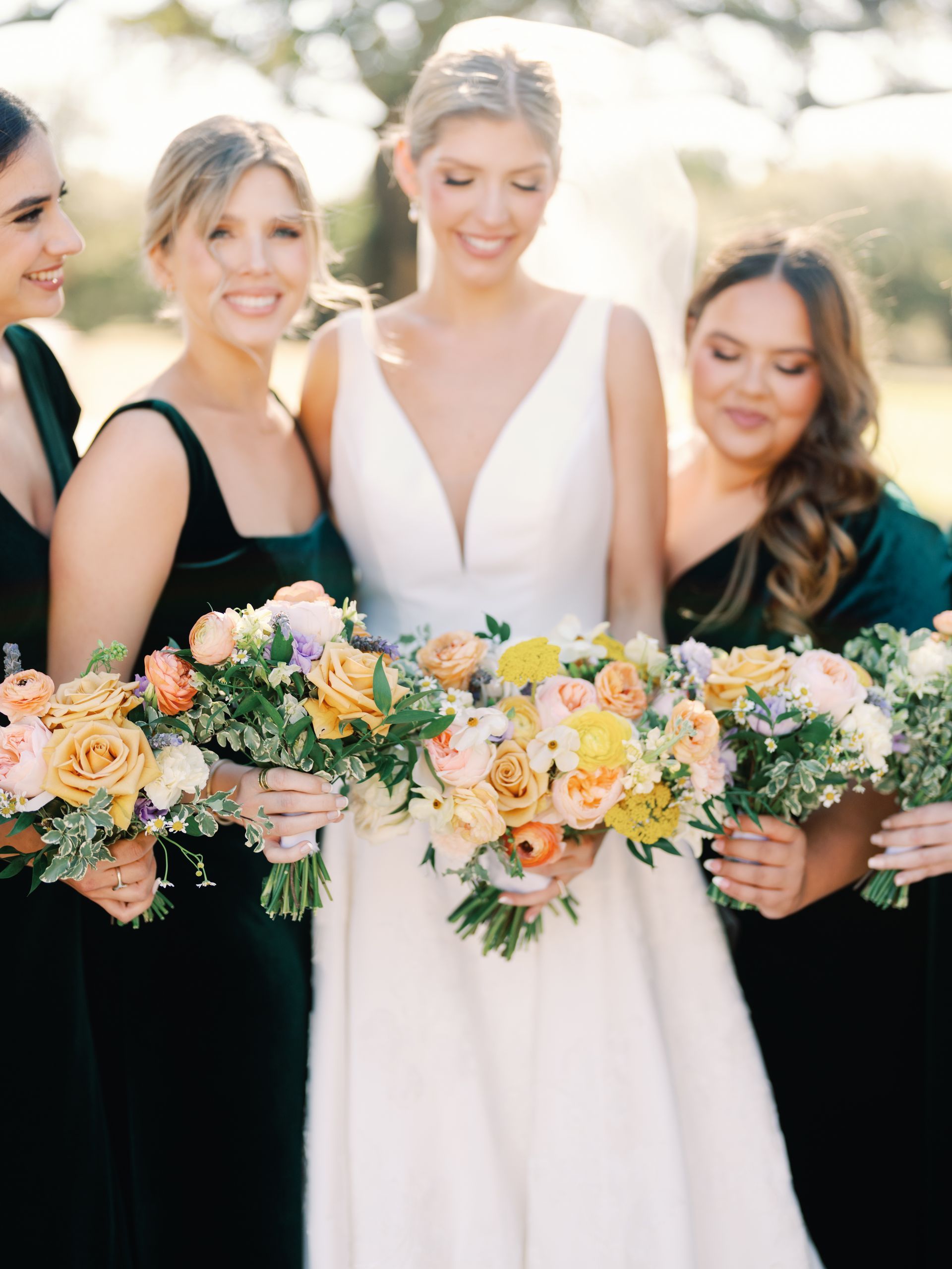 A bride in a white wedding dress stands with four bridesmaids in dark green velvet dresses, holding colorful bouquets.