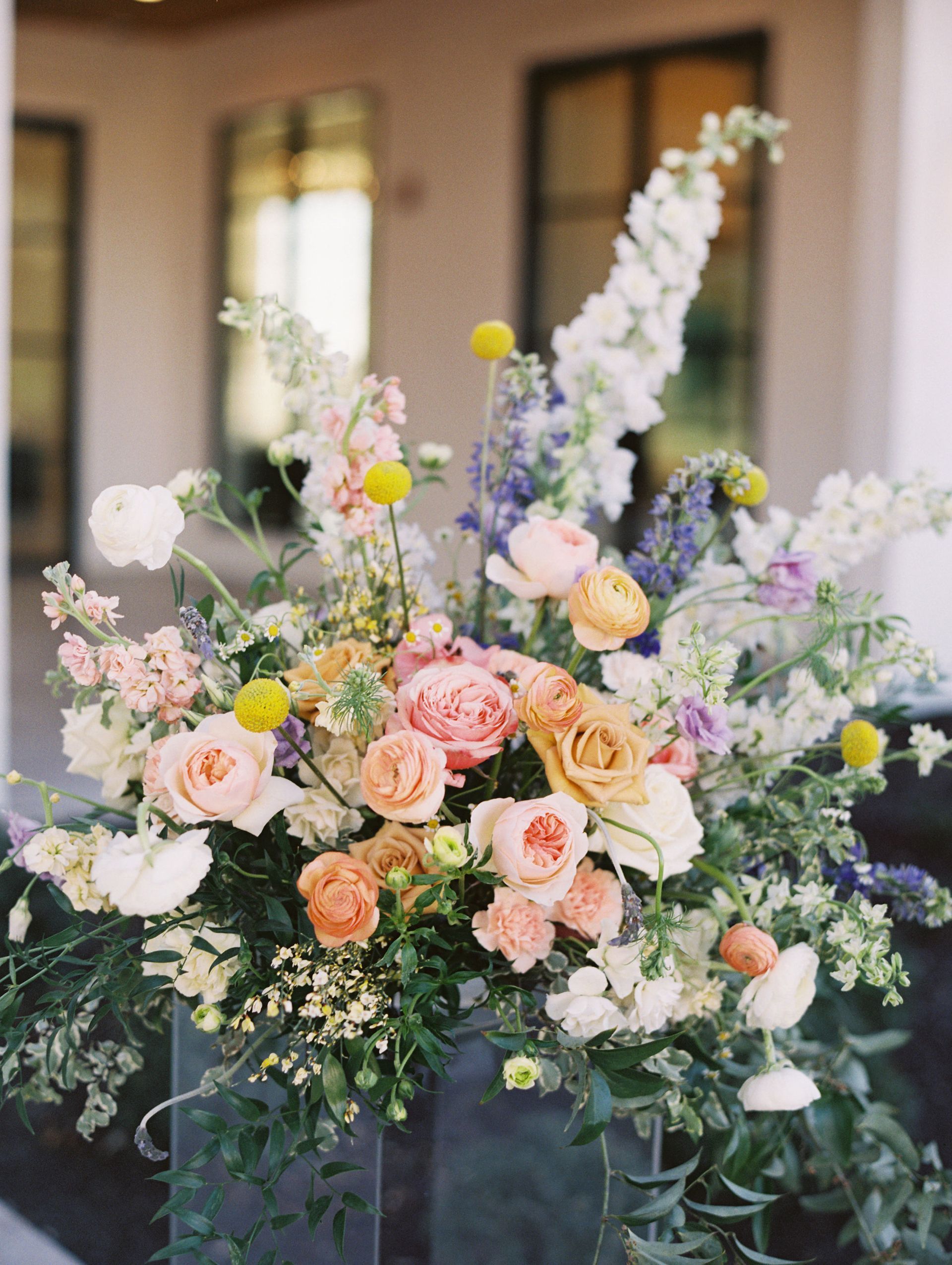 A large, lush floral arrangement featuring pastel roses, yellow craspedia, white delphinium, and greenery on a clear stand.