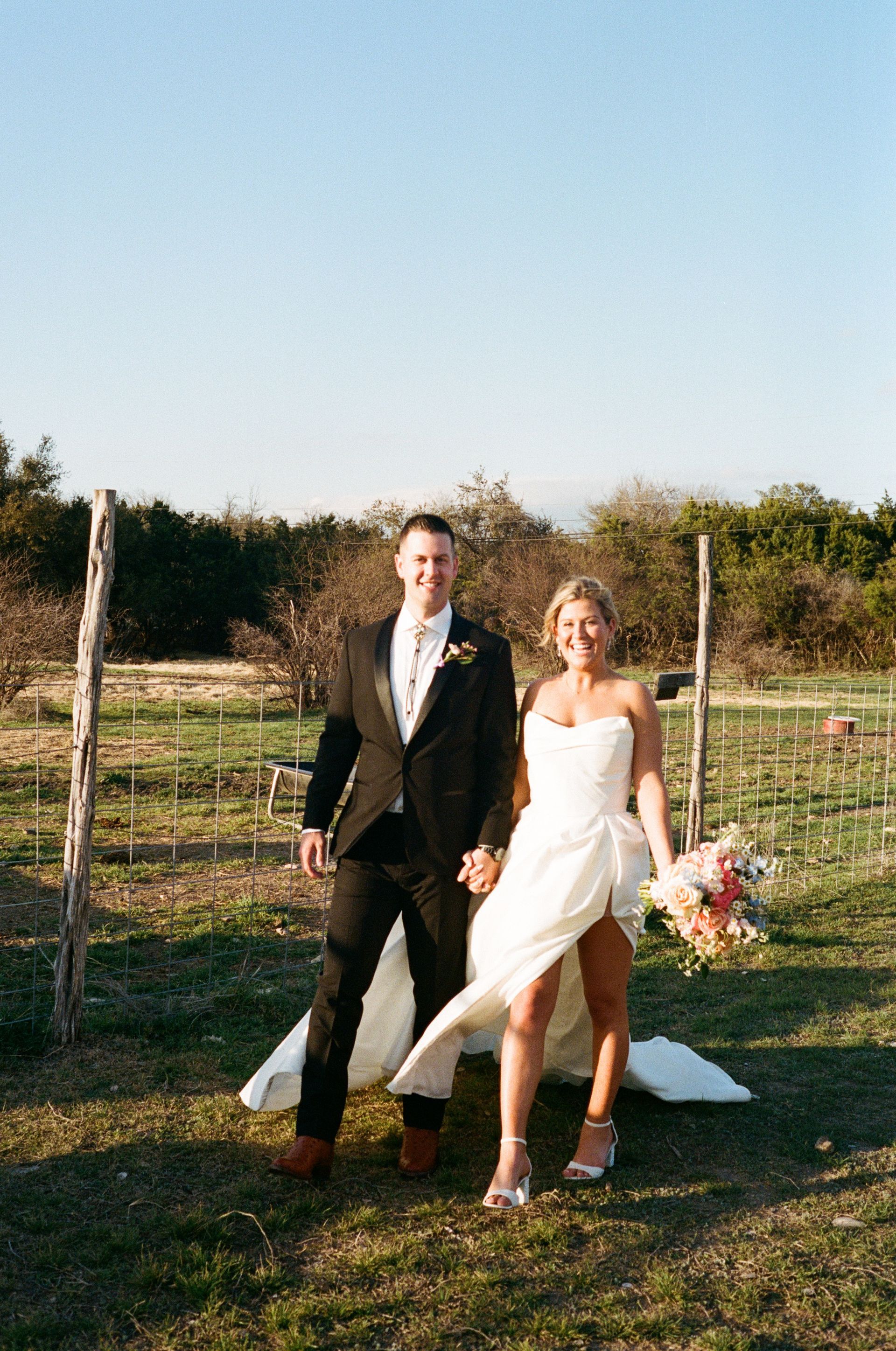 A newlywed couple holds hands while smiling in a sunny, rural outdoor setting.