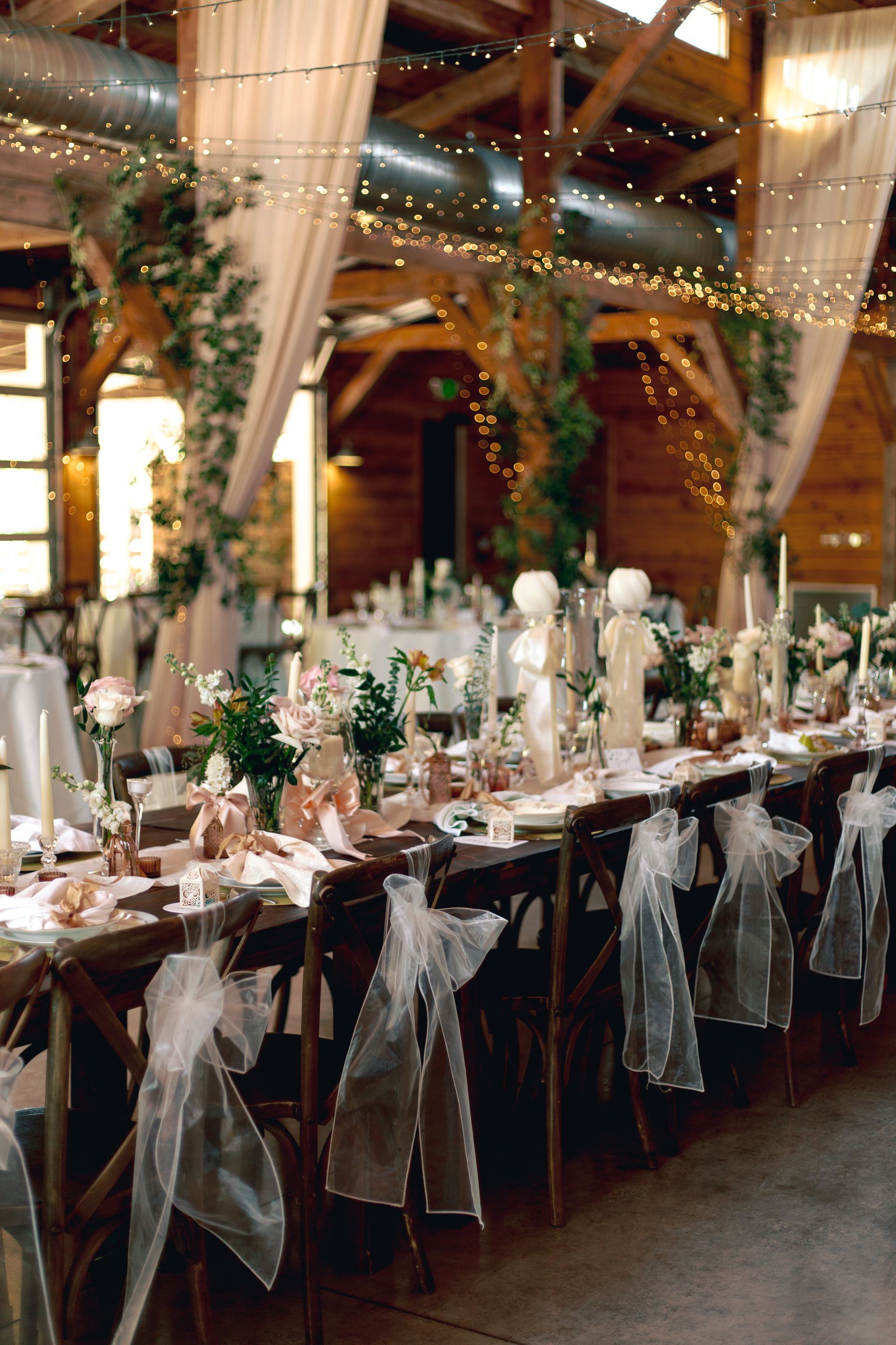 A rustic wedding reception table featuring elegant floral arrangements, white candles, and chairs draped with sheer fabric.
