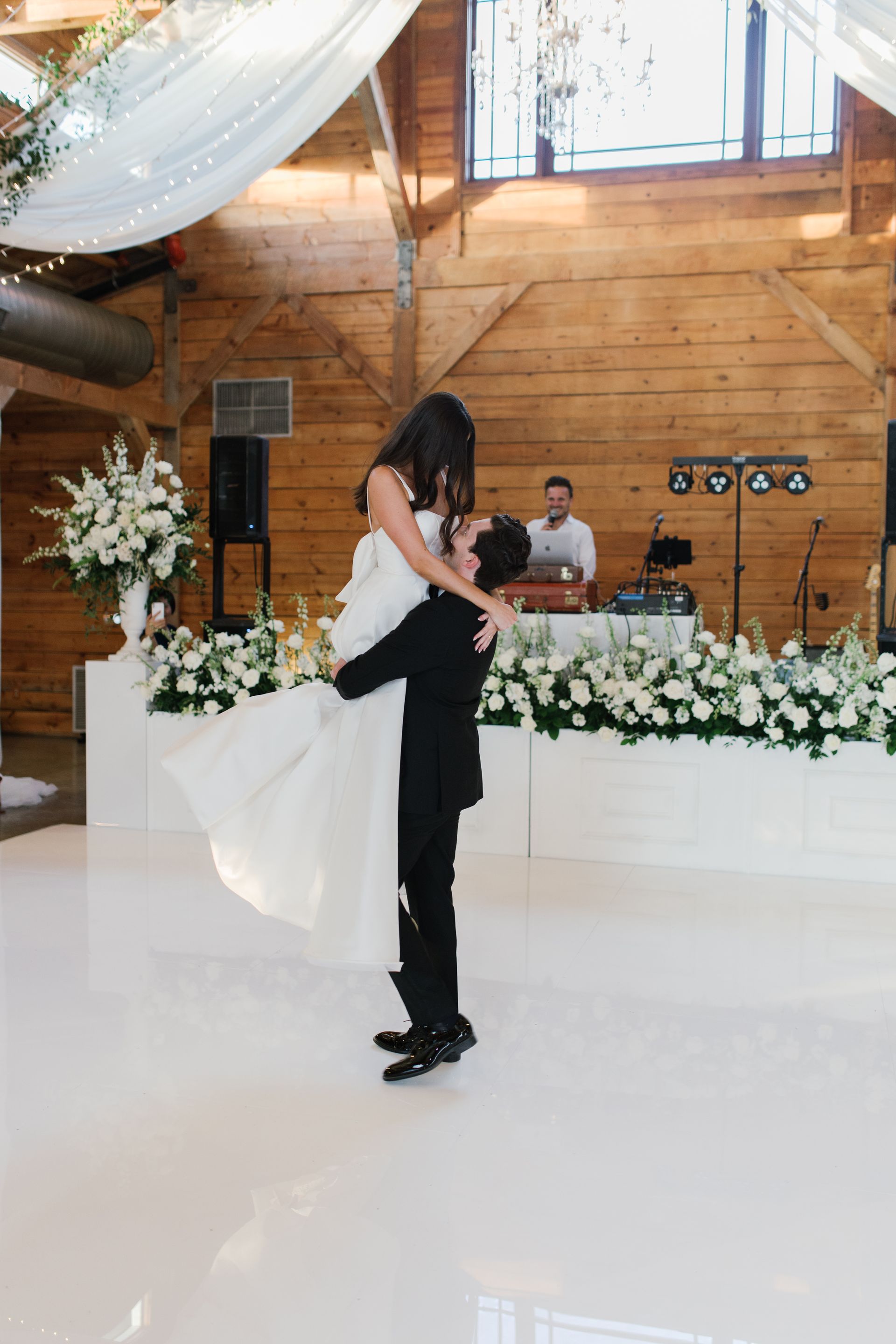 A person in a black suit lifts a person in a white wedding gown in their arms while dancing in a rustic indoor venue.
