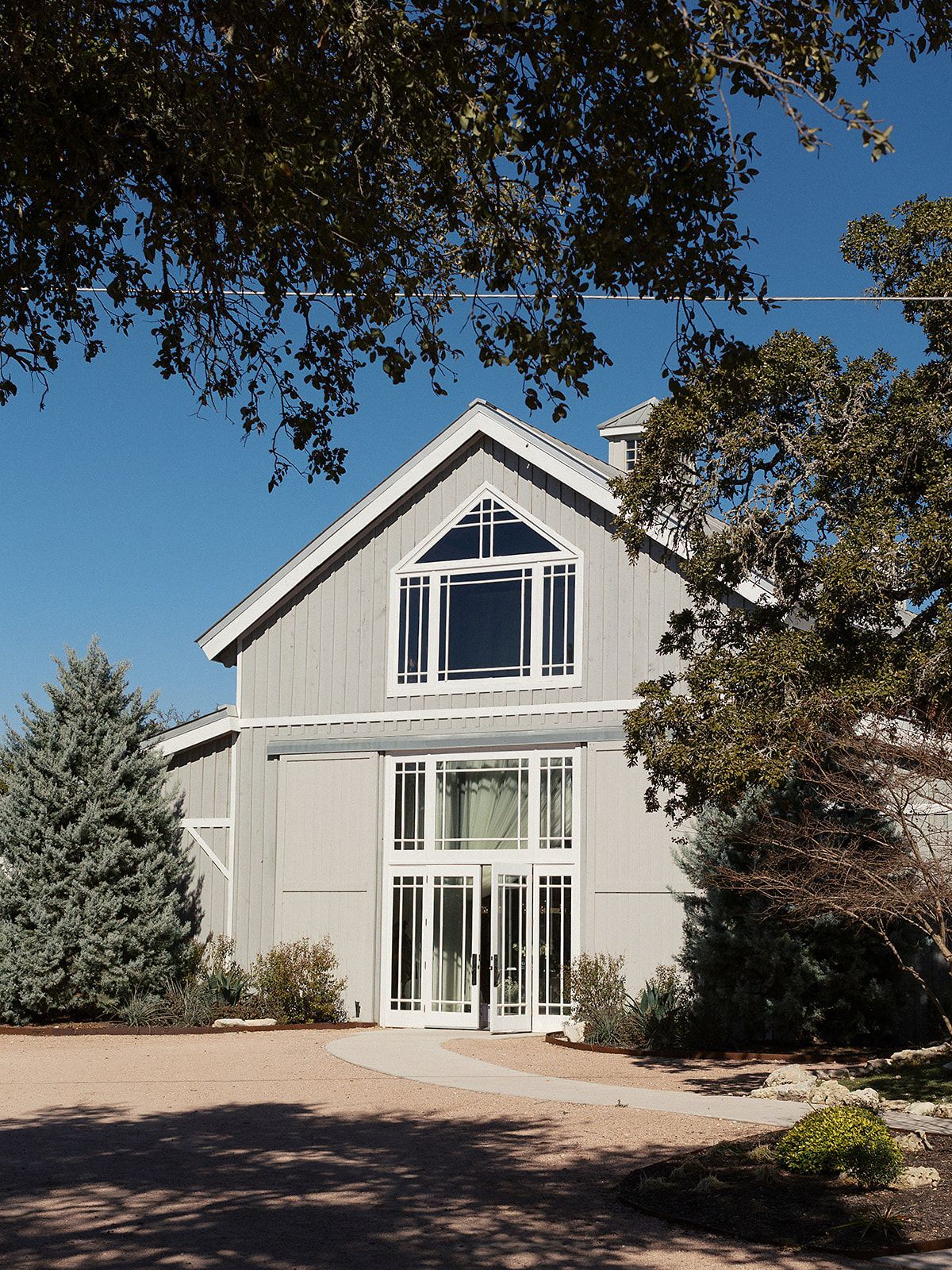 A light gray barn-style building with large glass doors and windows, framed by mature trees under a clear blue sky.