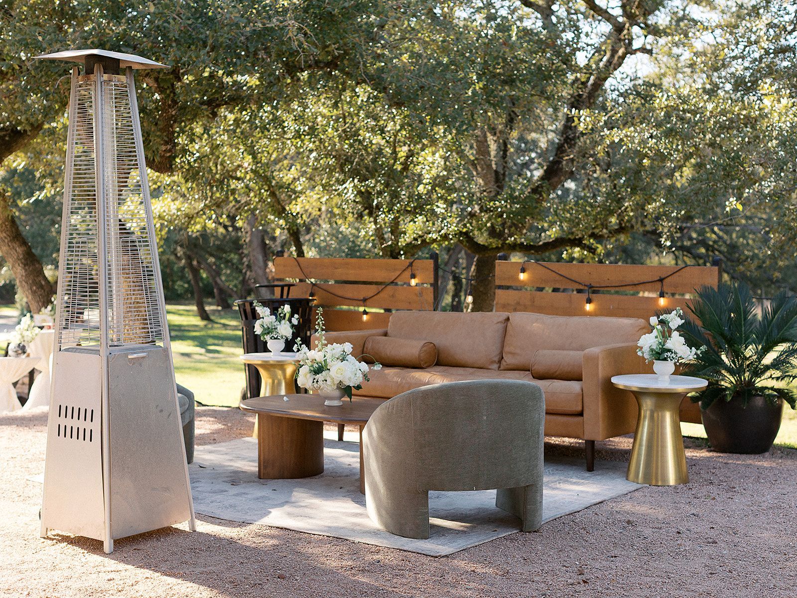 An outdoor seating area with a tan leather couch, a textured chair, and a patio heater under trees on a graveled lot.
