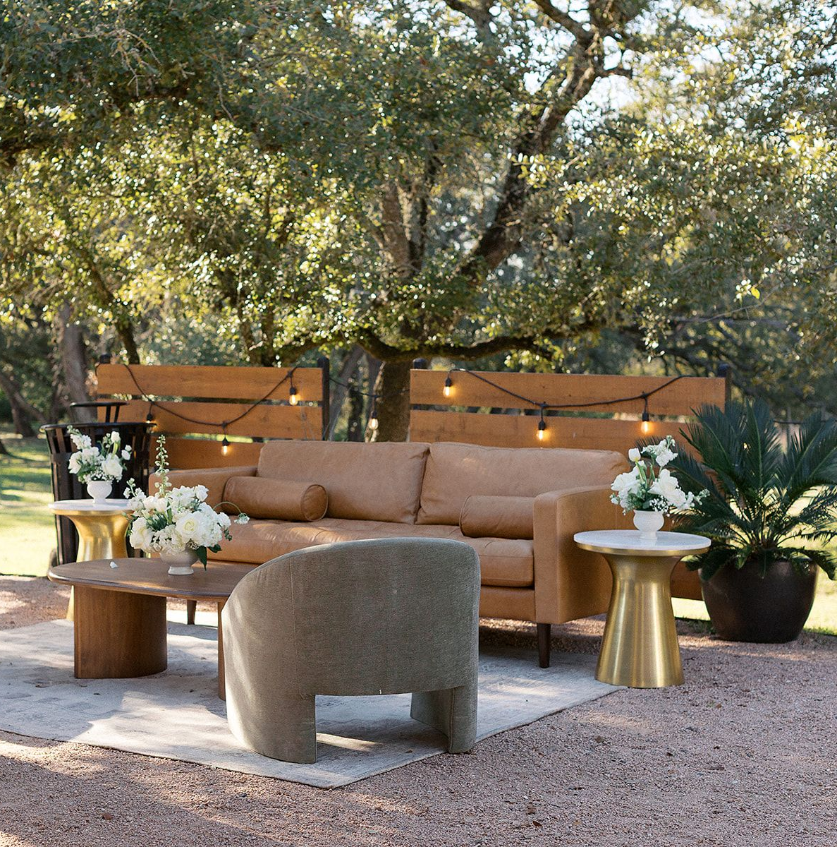 Outdoor lounge area with a tan sofa, rounded armchair, and two gold side tables on a light rug over gravel.